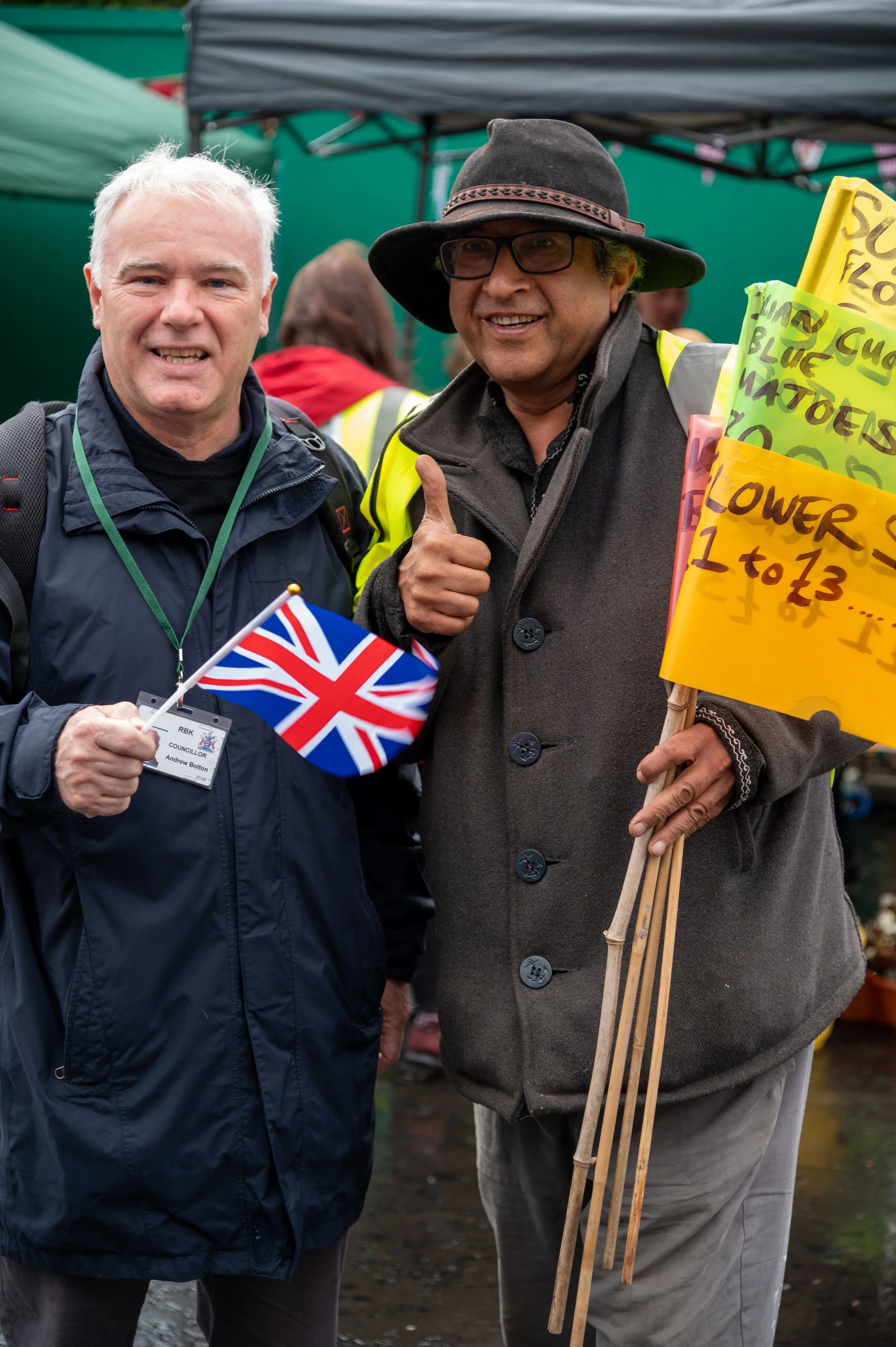 Two men smiling and standing outdoors at a protest or demonstration, holding a small Union Jack flag and a yellow sign with handwritten message, wearing jackets and hats, with tents and other people in the background.