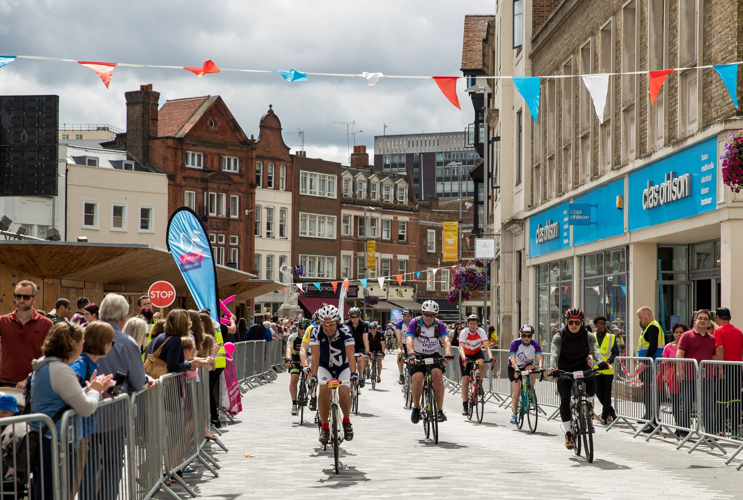Cyclists participating in a city street race, surrounded by spectators behind barriers, under gray sky with city buildings and banners overhead.