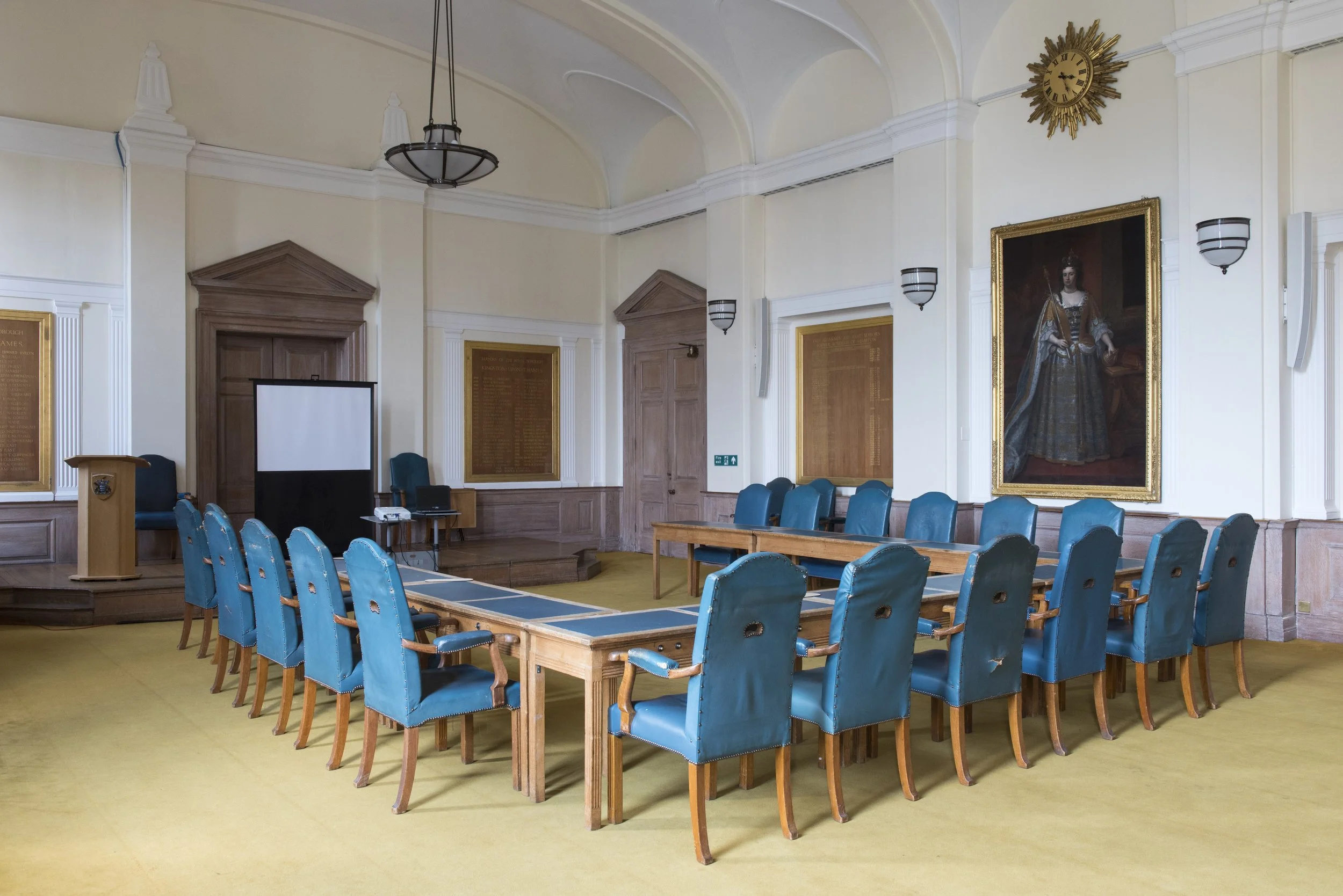 A large room with beige carpet and cream-colored walls, furnished with a long wooden table surrounded by blue upholstered chairs, a wooden podium, a projection screen, and framed portraits on the walls, including a master portrait of a woman in historical attire, and a clock on the wall.
