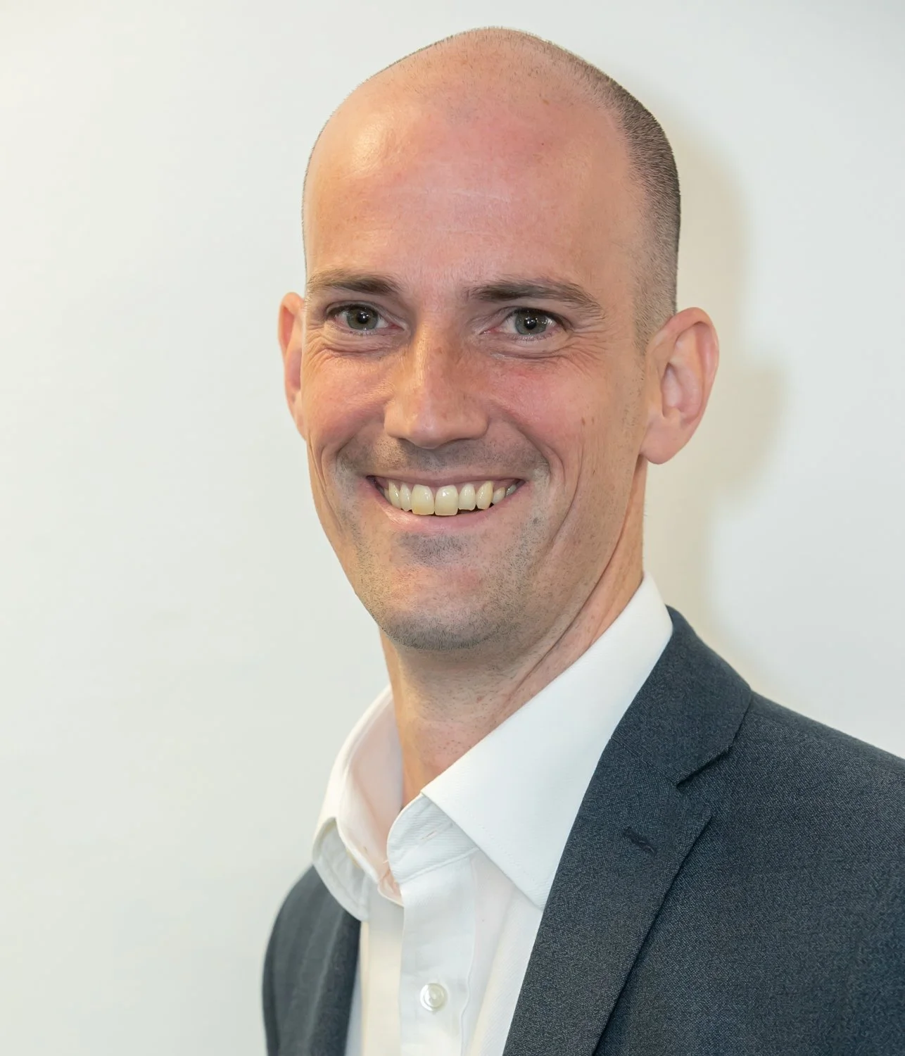 A smiling man with a shaved head wearing a white shirt and dark suit jacket, standing against a plain white wall.