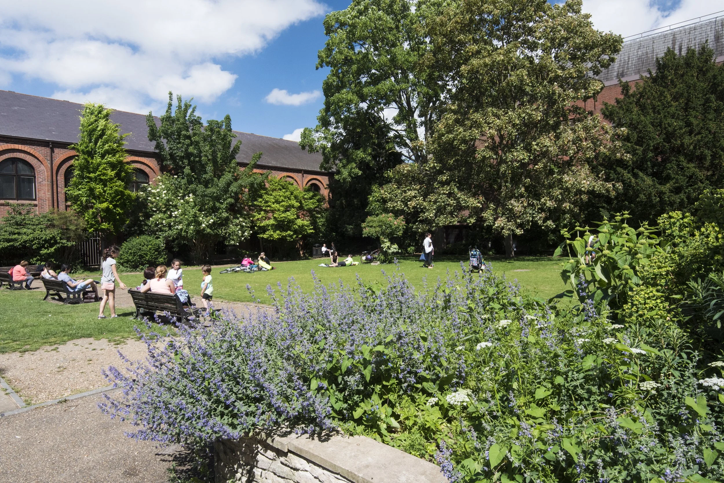 A park scene with children and adults sitting and walking on the grass and benches, surrounded by trees and purple flowers, with a brick building and blue sky with clouds in the background.
