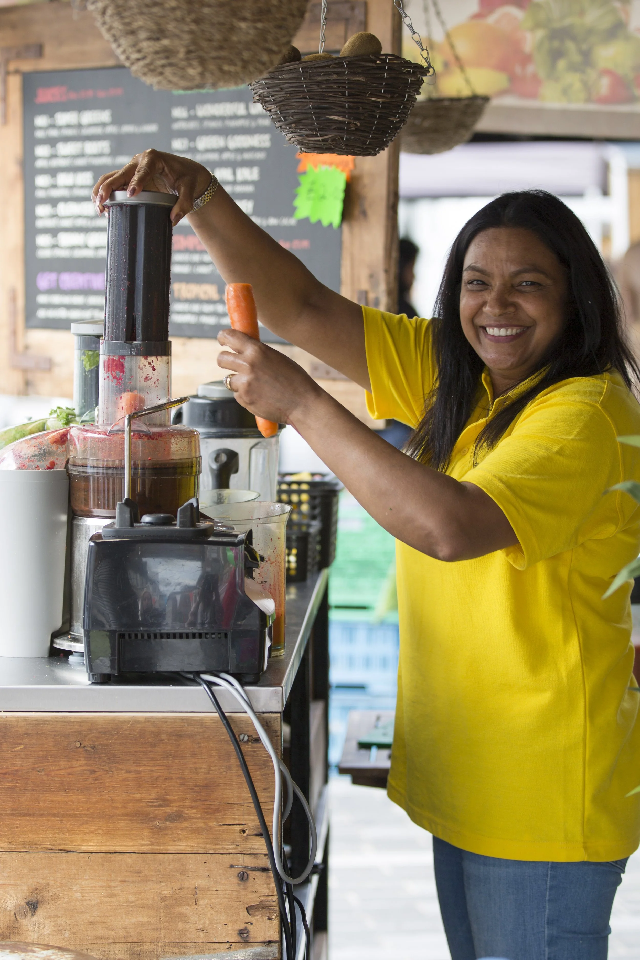 A woman in a yellow shirt is smiling while using a juicer machine at a market stand, holding a carrot in her right hand and operating the juicer with her left hand. The stand has a wooden counter and hanging baskets above.