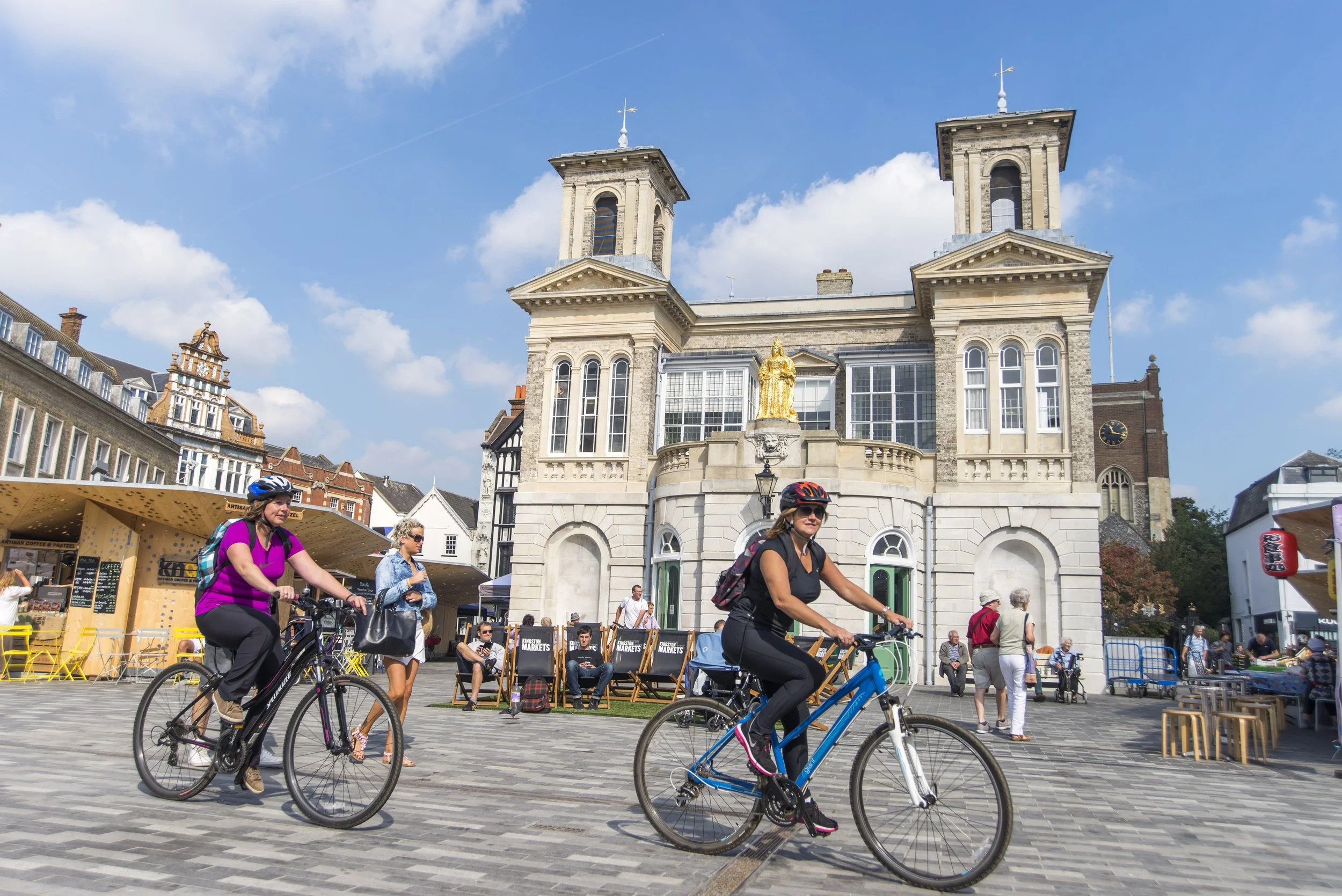 People riding bicycles and walking in a busy town square with historic buildings, outdoor markets, and a bright blue sky.