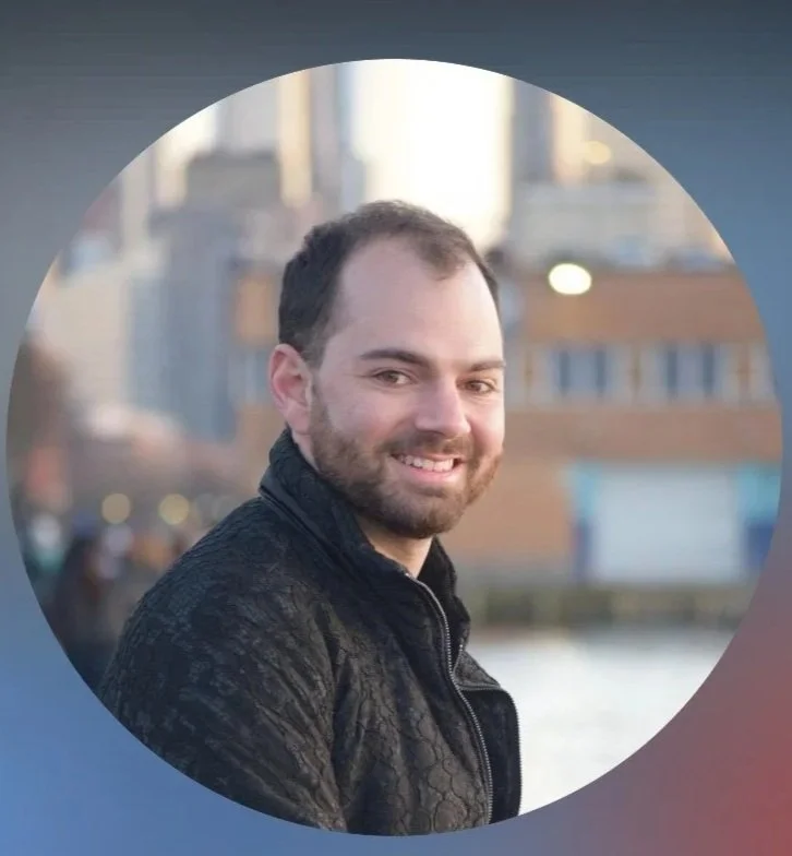 A smiling man with a beard and short hair, wearing a black textured jacket, standing outdoors near a body of water with buildings in the background.