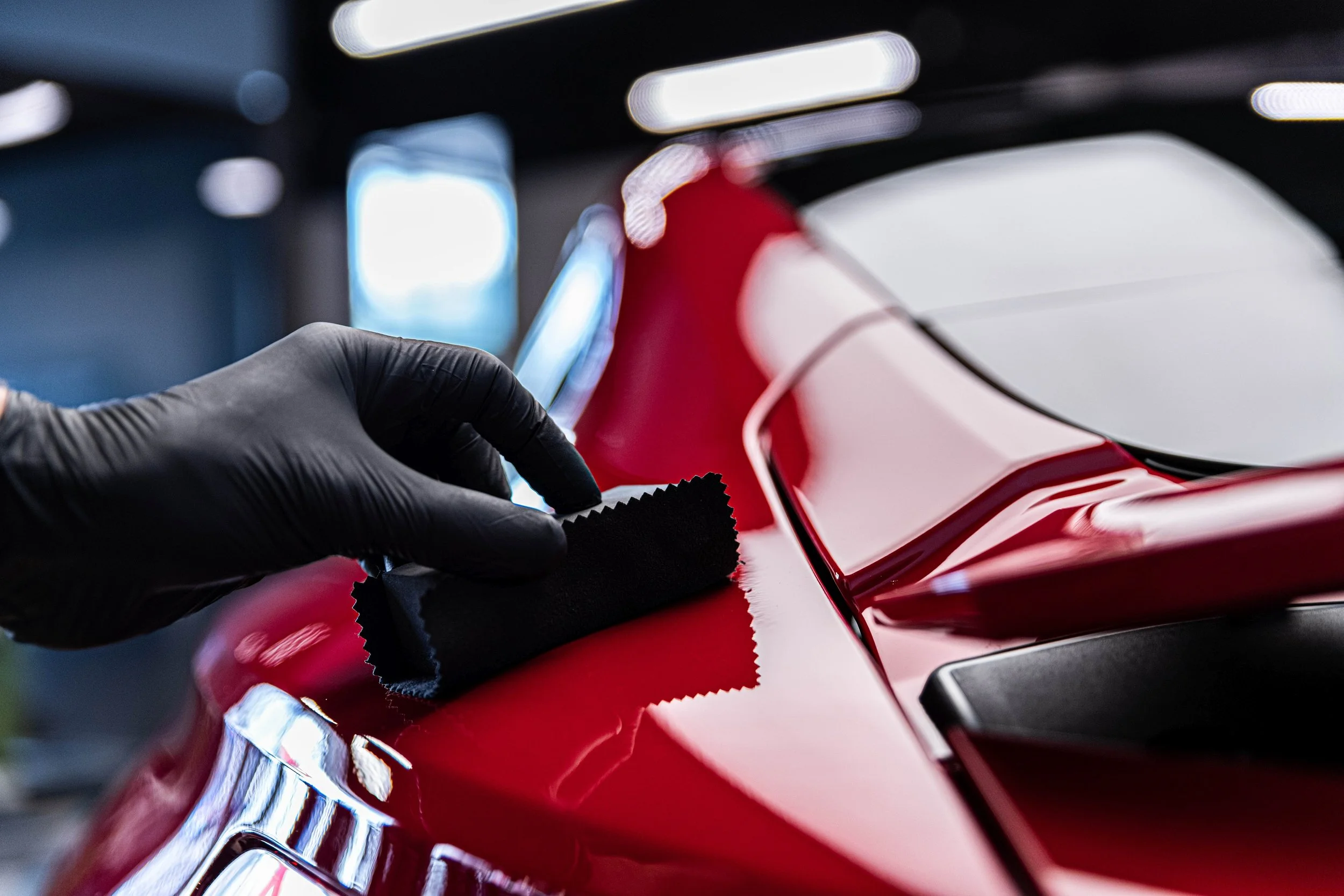 A person wearing black gloves polishing a shiny red car with a pad in an auto detailing setting.