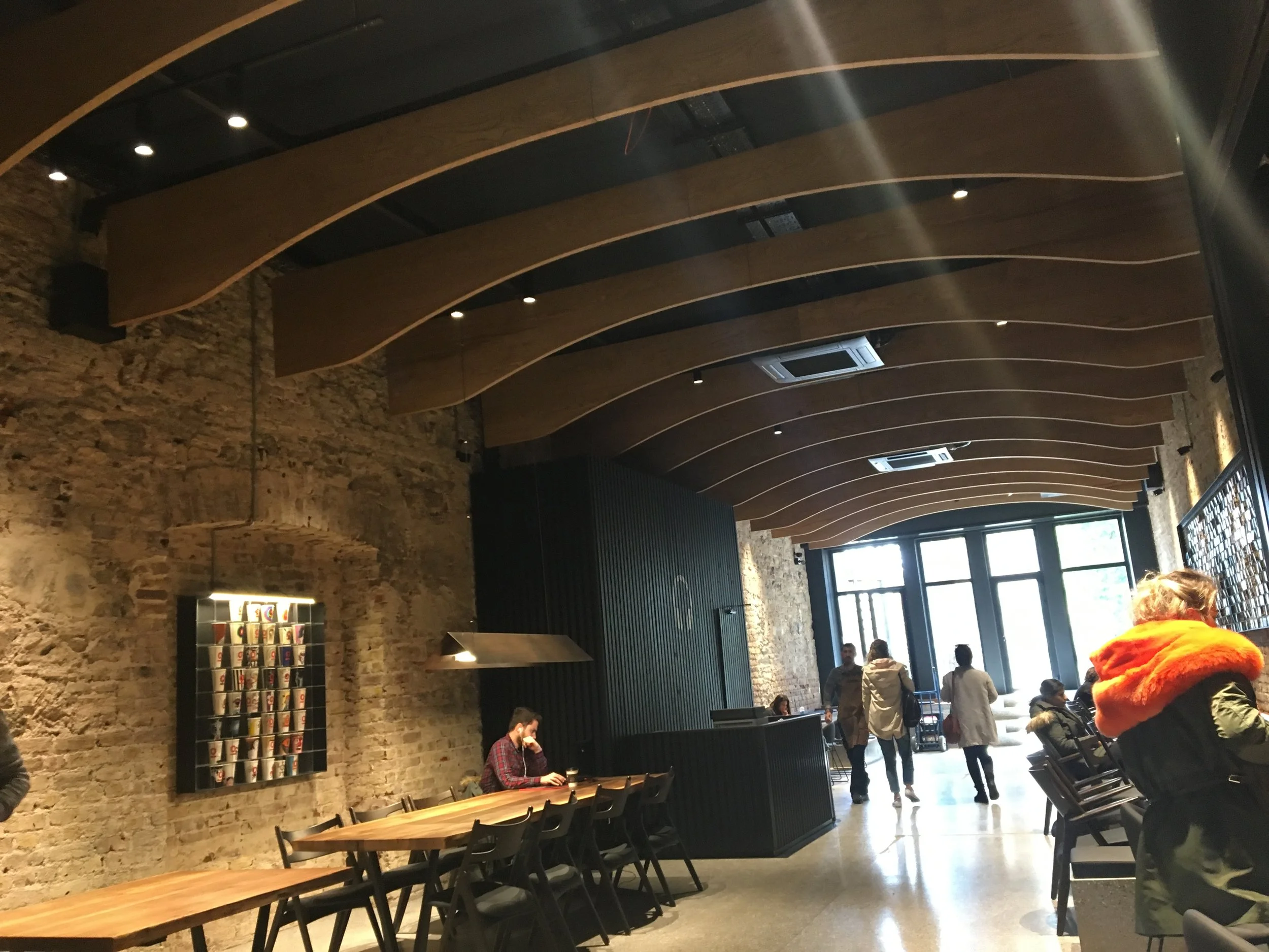 Modern ve şık bir kafenin iç mekanı, ahşap kirişler ve taş duvar dikkati çekiyor. Tavandan sarkan lambalarla aydınlatılmış, uzun ahşap masalar ve sandalyeler bulunuyor. Arka planda duvarda kahve fincanlarının sergilendiği bir raf mevcut. Çeşitli insanlar oturmuş, içeri girip çıkıyor.