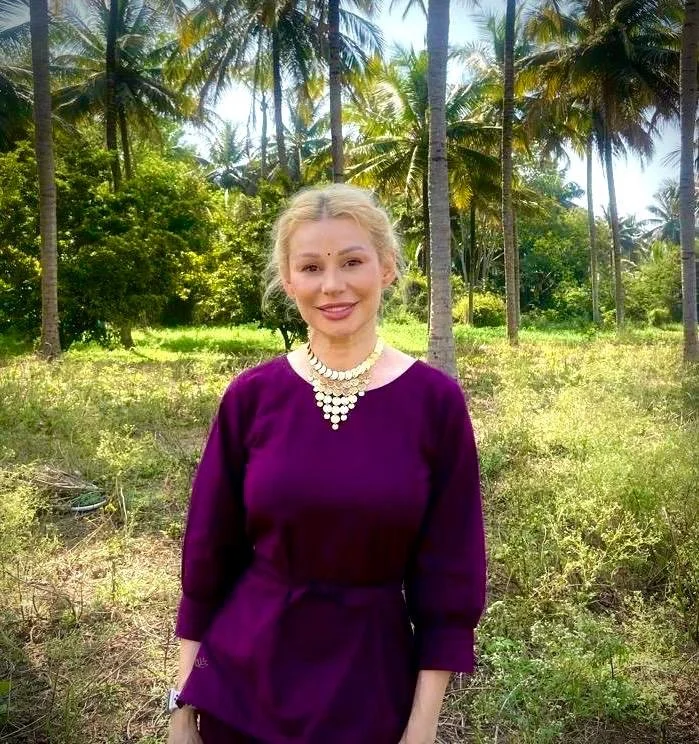 A woman with blonde hair in a purple dress and pearl necklace smiling outdoors in a tropical setting with palm trees and green foliage in the background.