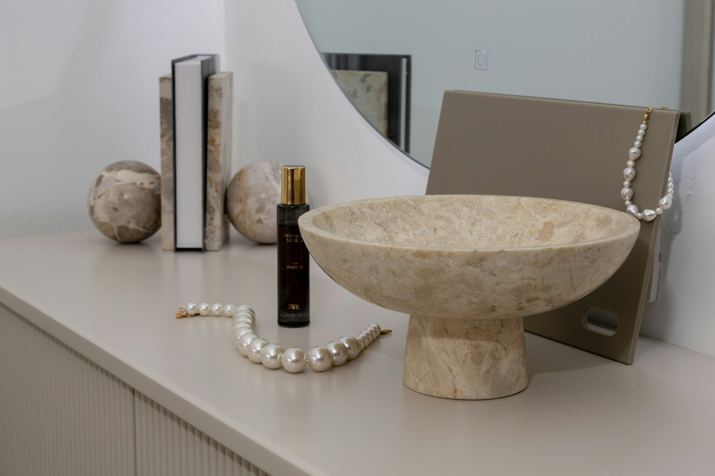 Decorative marble bowl with jewelry, books, perfume bottle, and rocks on a white surface in front of a mirror.