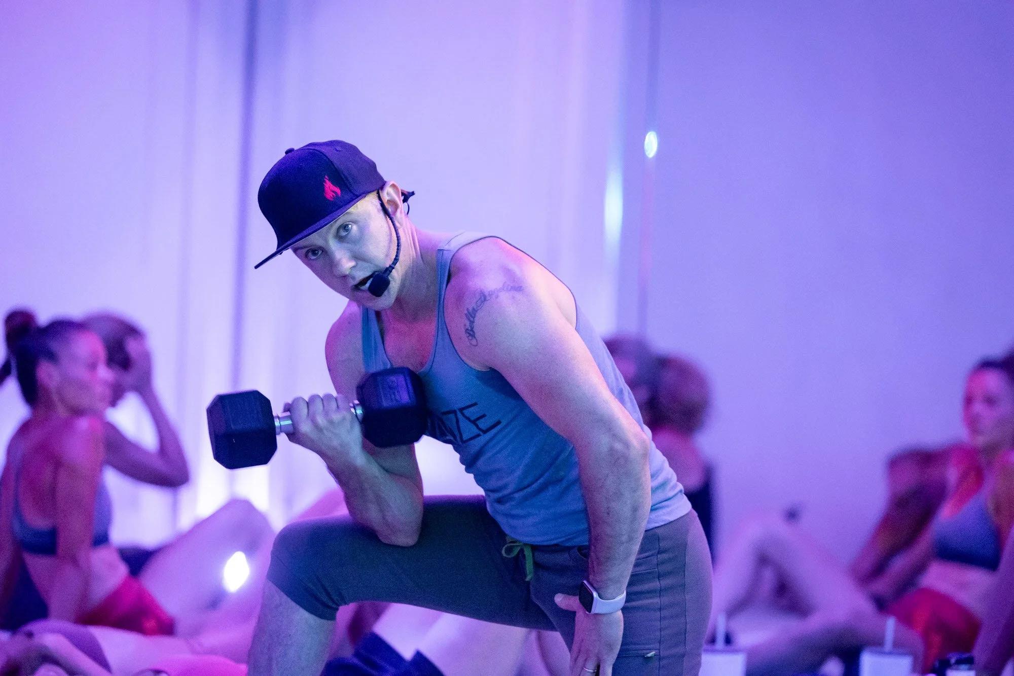 image of a fit man in a baseball cap with a flame on it in a blue tank demonstrating a Concentration Curl in a fitness class