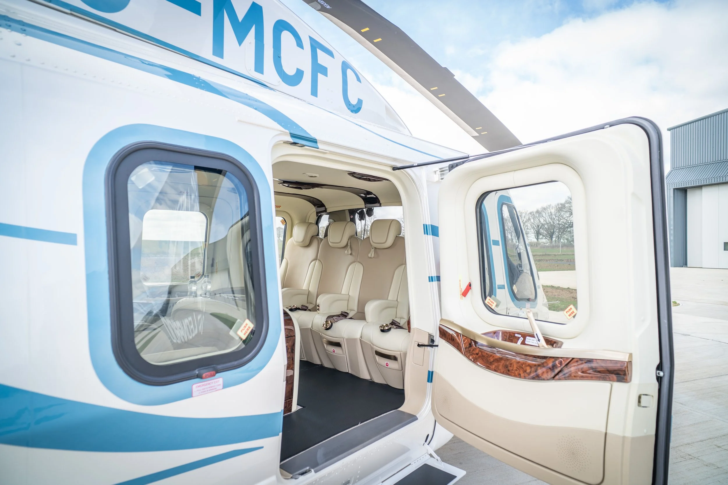 Interior of a medical helicopter with beige seats and large windows, outside view of a gray industrial building and cloudy sky.