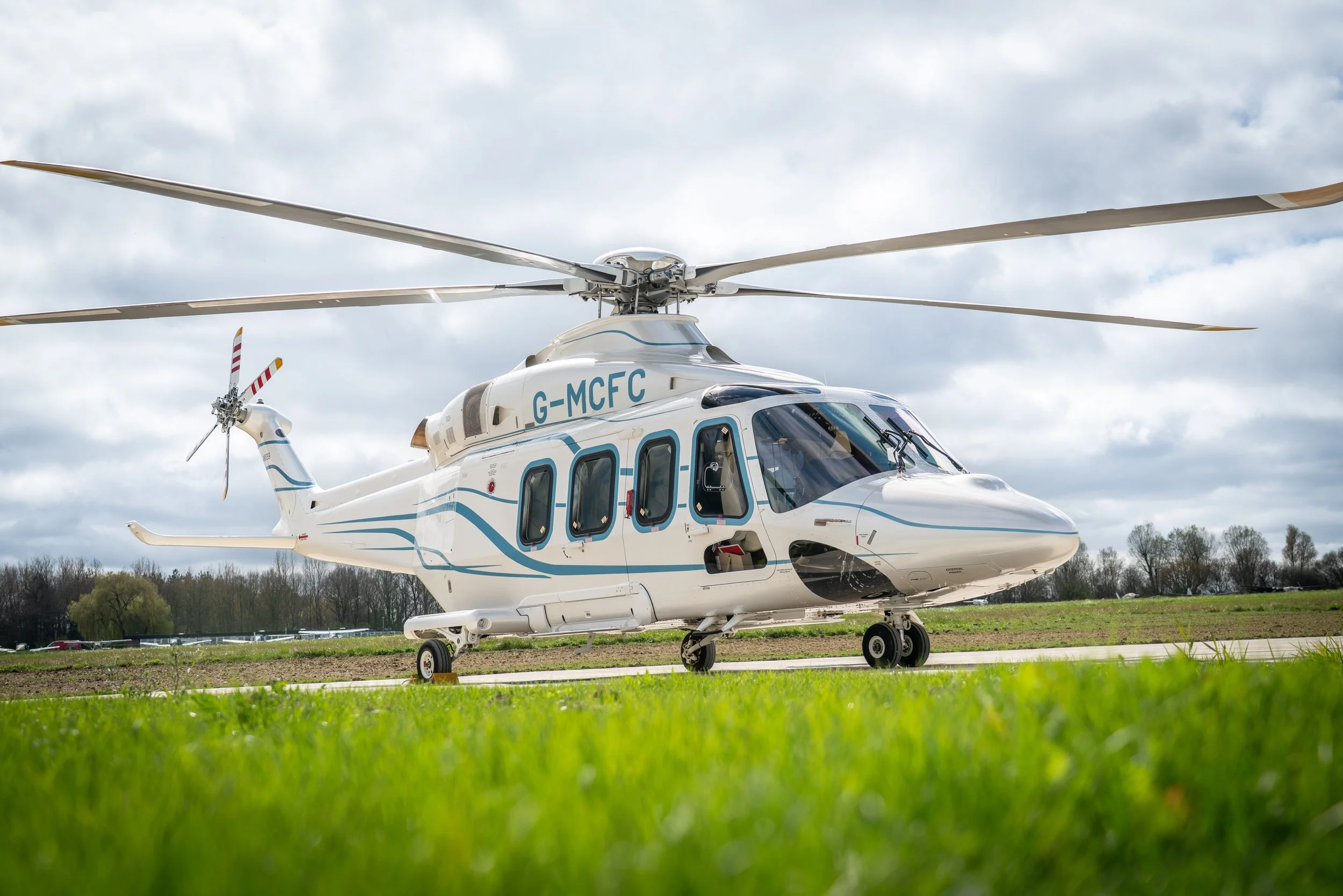 A white helicopter with blue accents parked on grass, with cloudy sky background.