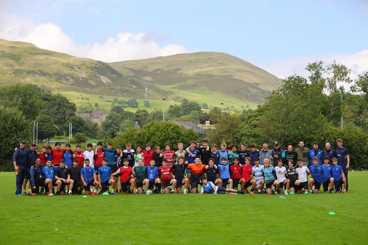 Group photo of young rugby players and coaches on a grassy field with green hills in the background.