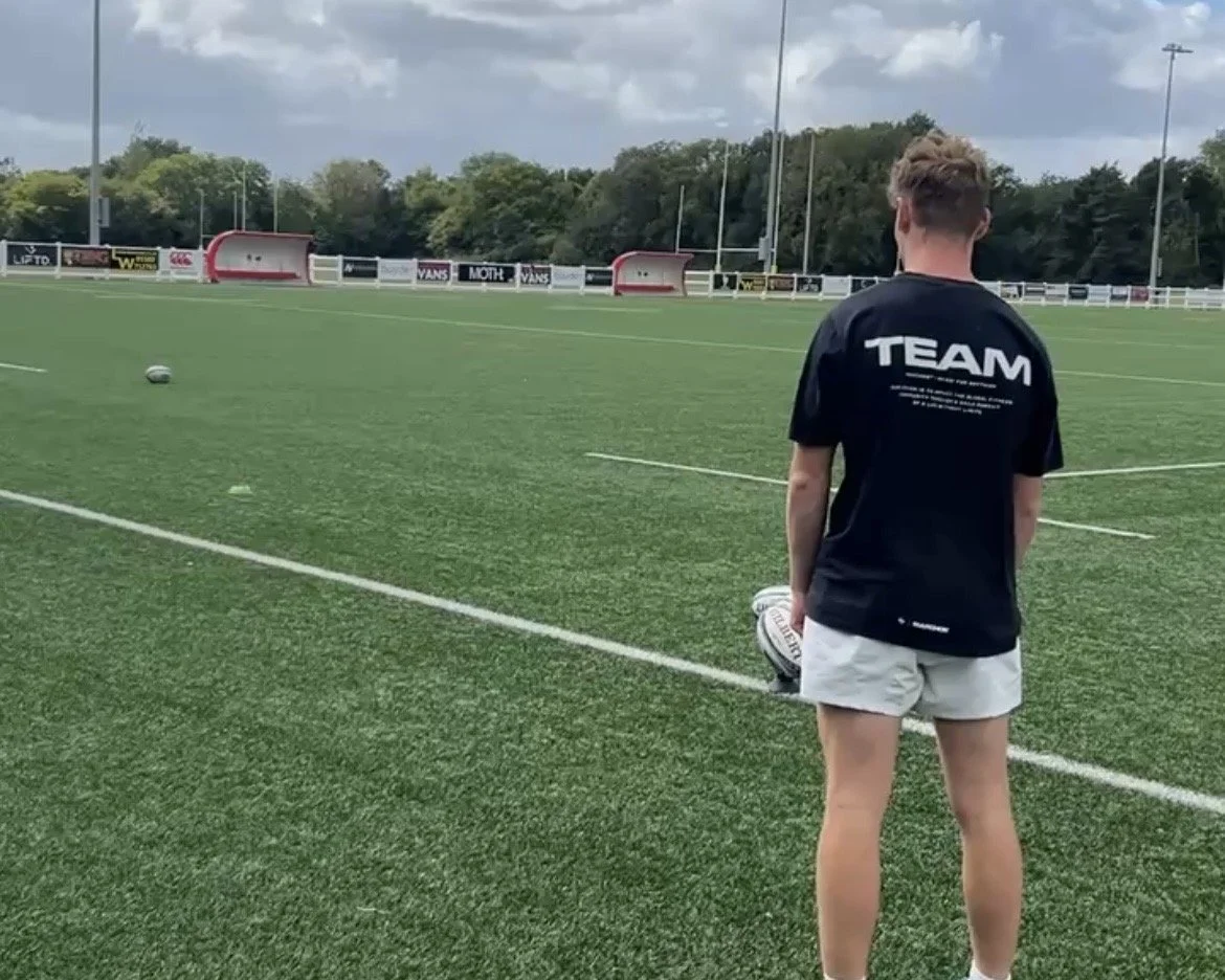 A person standing on a rugby field holding a rugby ball, wearing a black T-shirt with "TEAM" written on the back, and shorts.