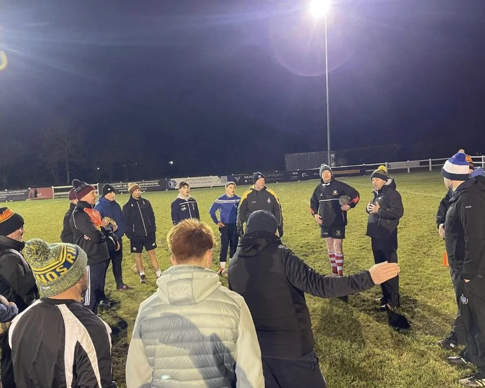 A group of rugby players and coaches gathered on a field at night, listening to a coach during a team meeting or strategy discussion, with bright stadium lights overhead.