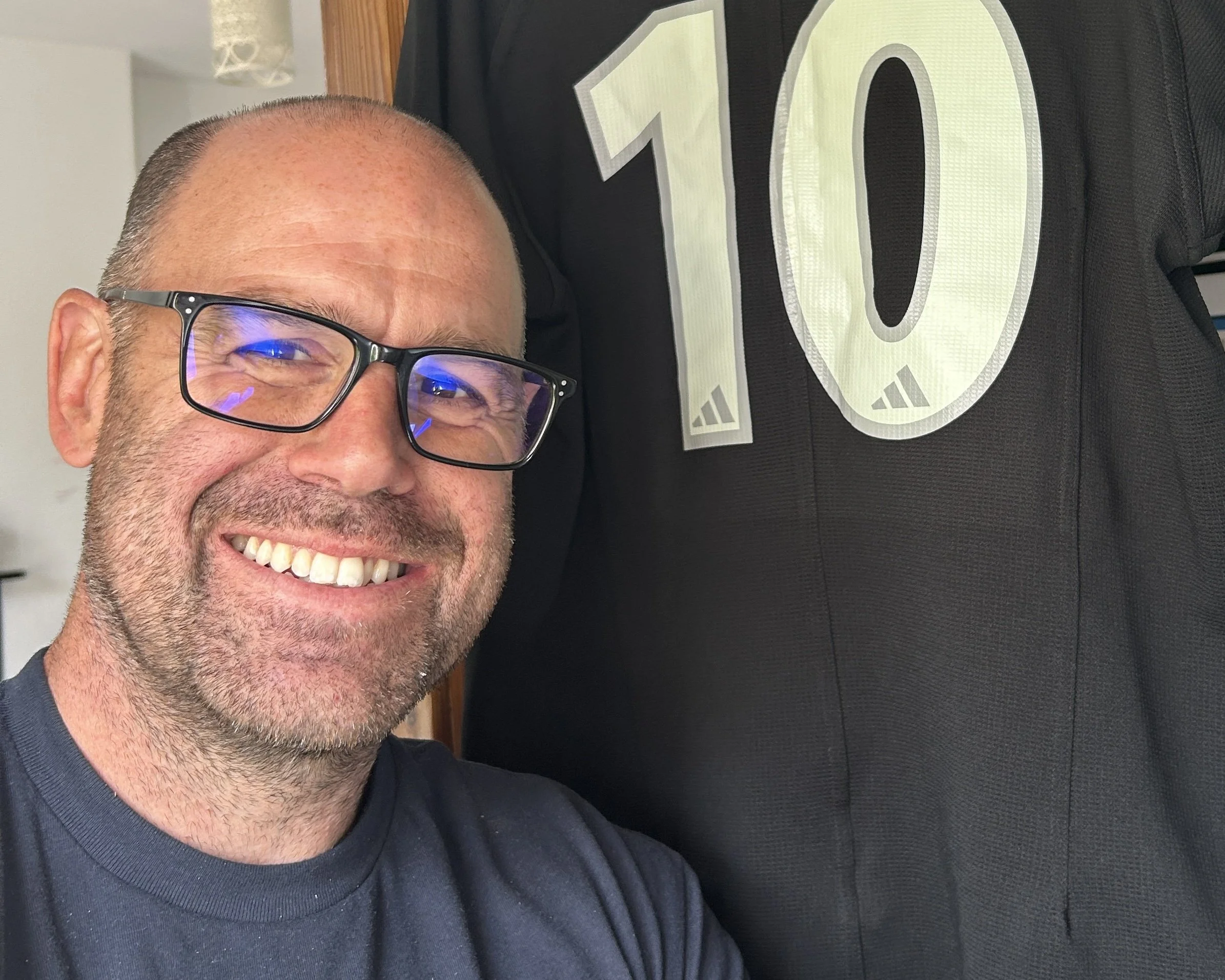 A smiling man with glasses takes a selfie next to a black jersey with the number 10 on it. The man has short hair and a beard.