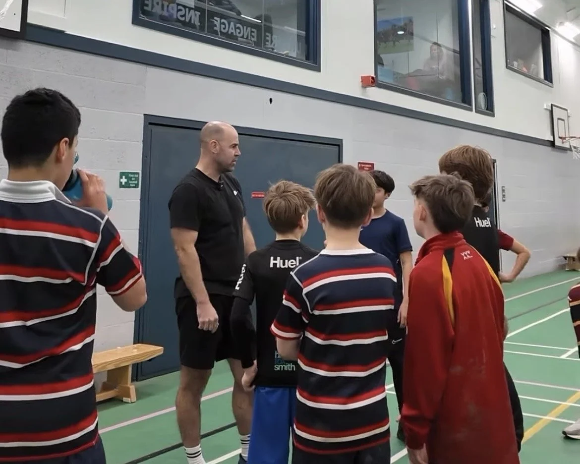 A group of young boys gathered around an adult male coach on a sports court, with additional people visible through a window above.