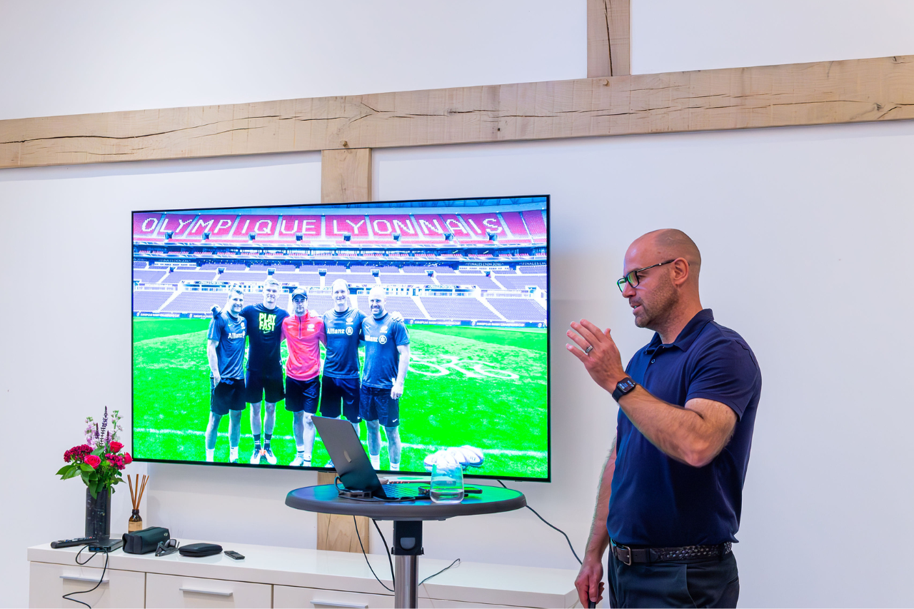 A man with glasses and a blue polo shirt standing beside a large screen displaying a photo of four soccer players on a field inside a stadium.