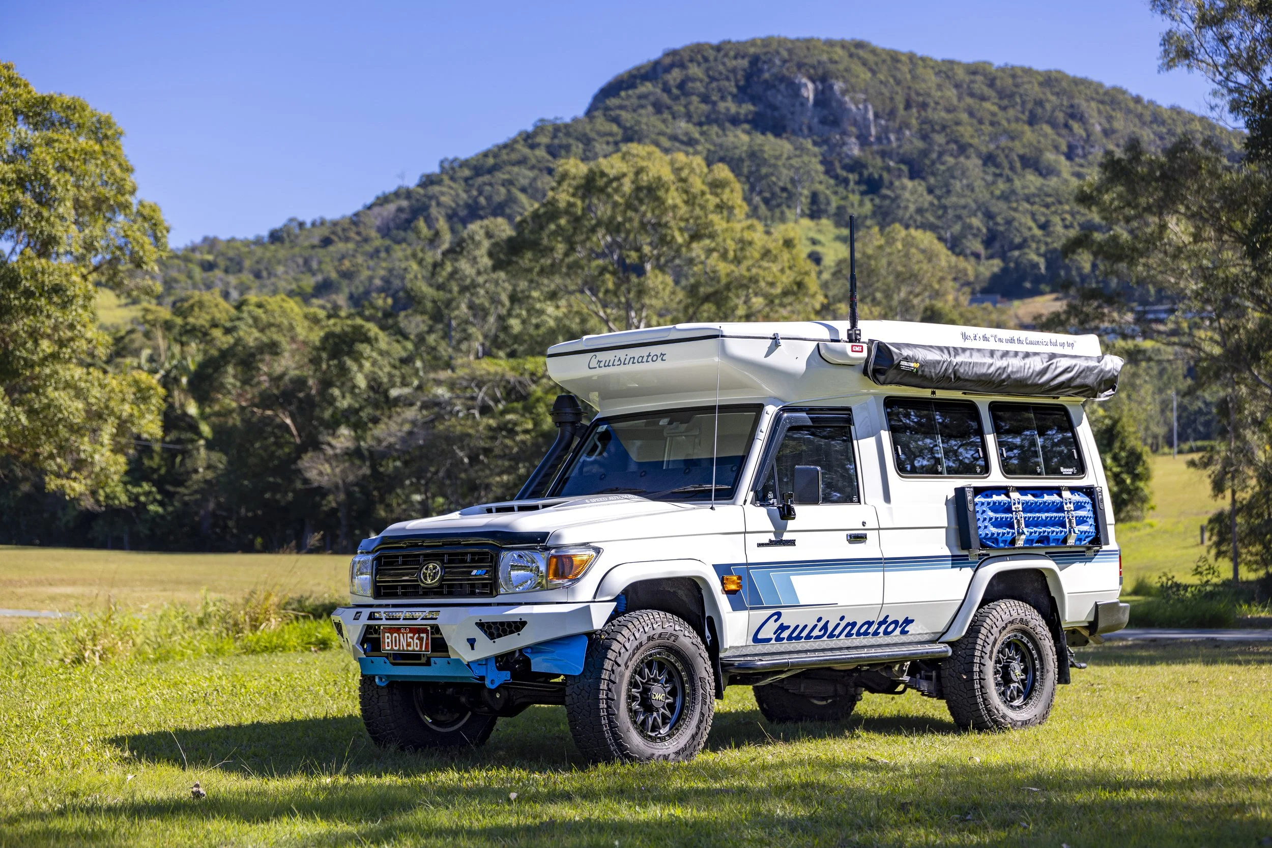 White Toyota Land Cruiser camper conversion with a rooftop tent, labeled 'Cruisinator,' parked on grass with a mountain in the background.