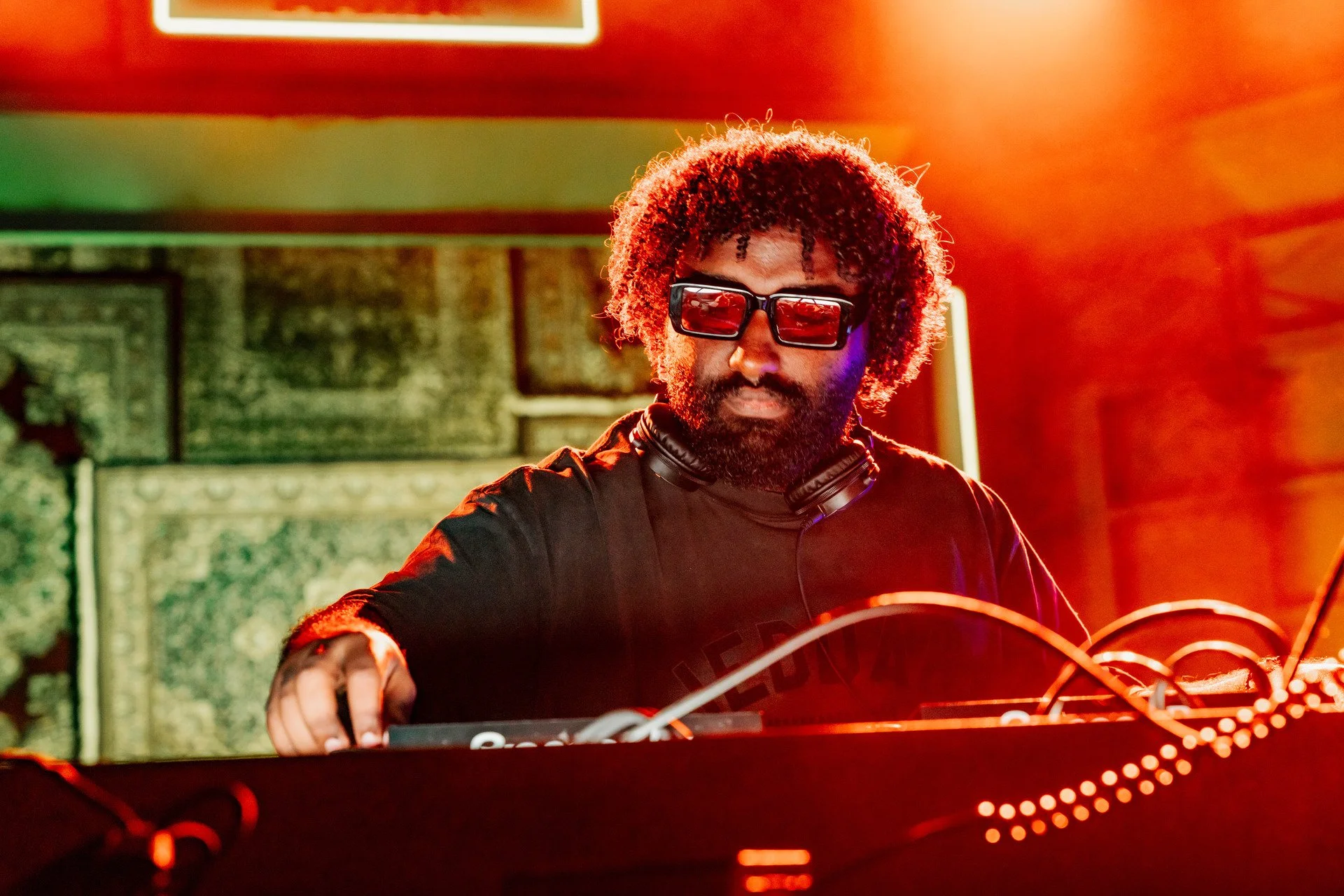 A DJ with curly hair, beard, wearing sunglasses and headphones around his neck, performing at a club with colorful lighting.