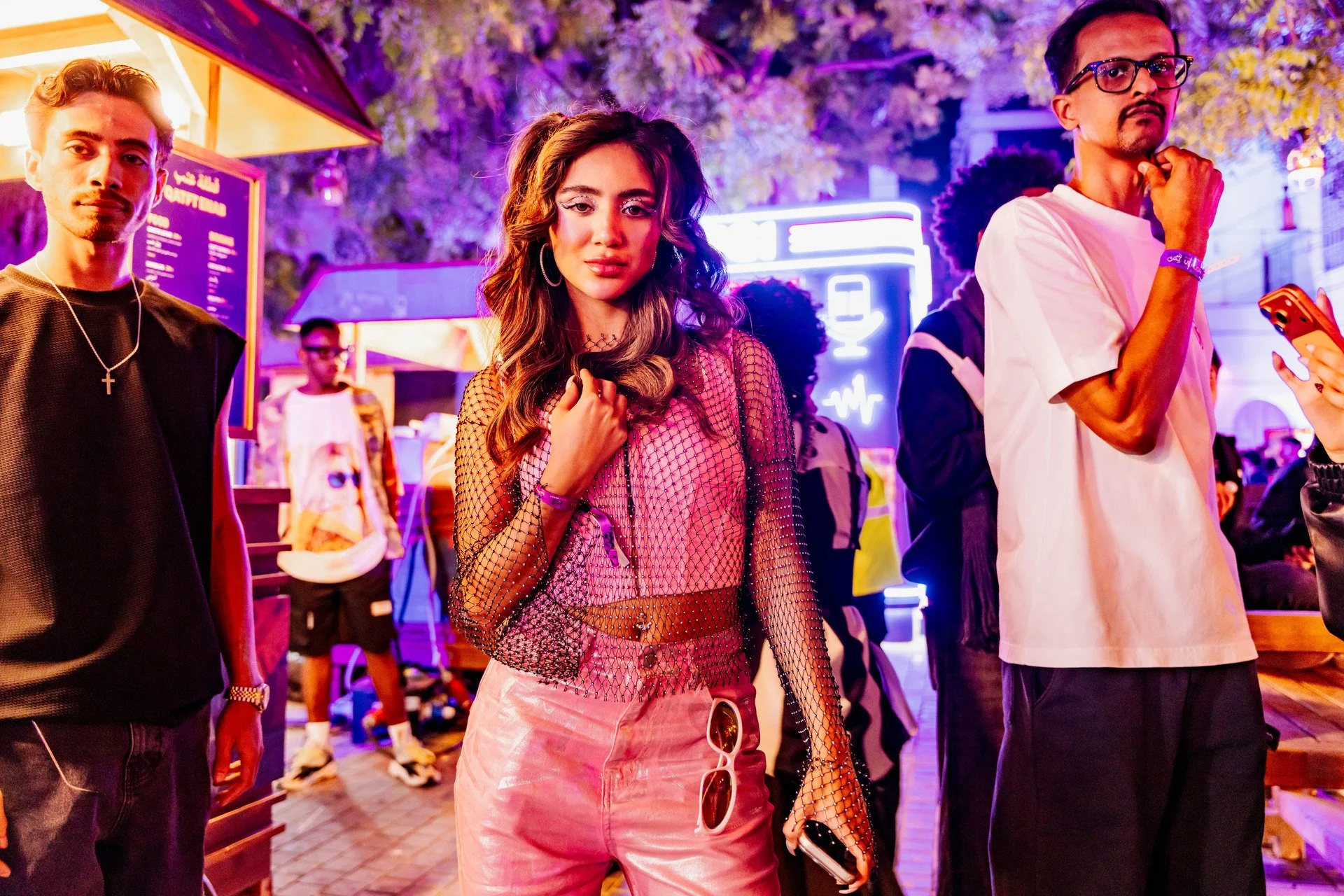 Young woman in pink mesh top and pants holding a phone at night market with neon lights, surrounded by young men, one with glasses and another with a cross necklace, under purple and yellow lights.
