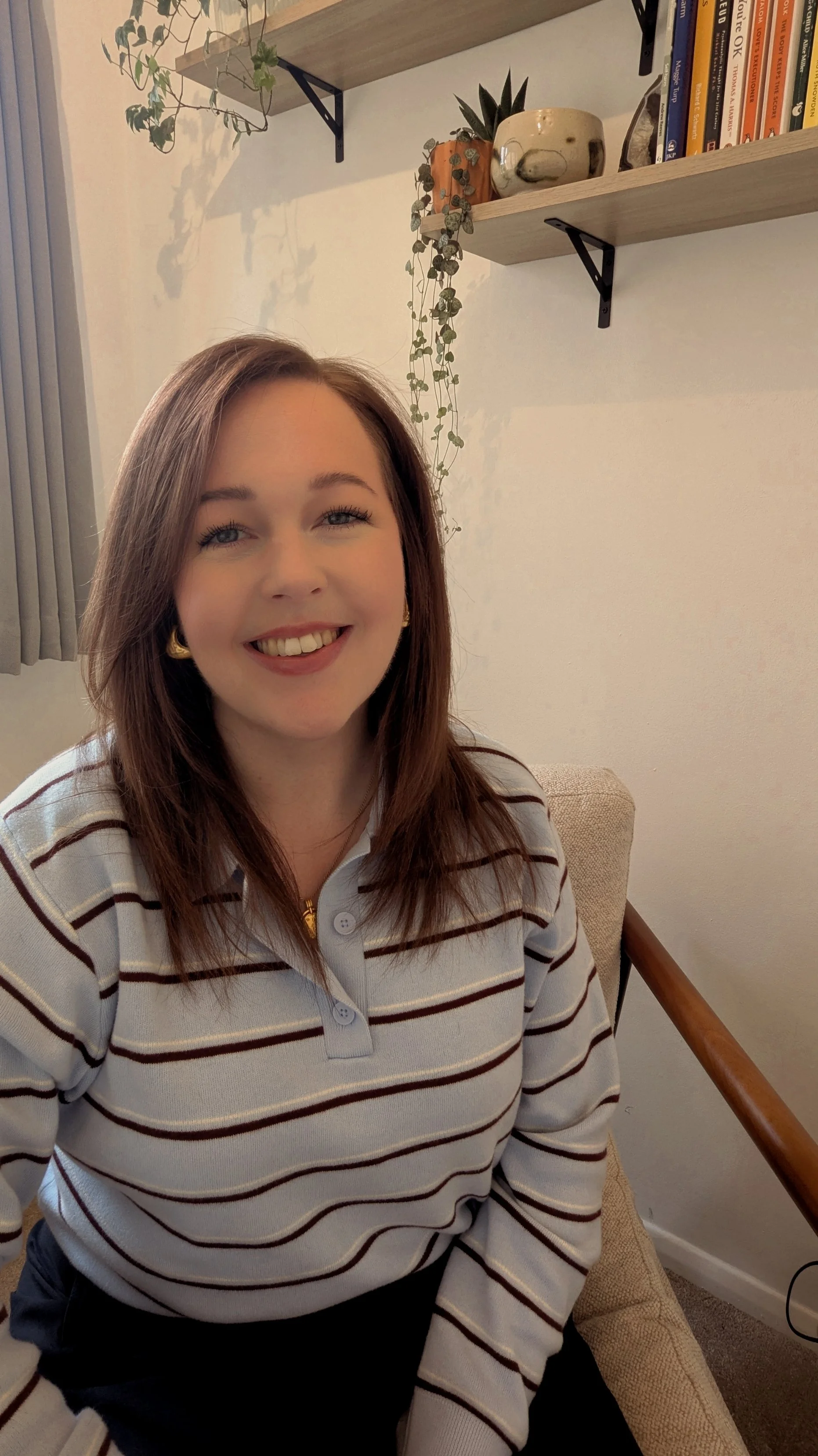 A woman in a cozy sweater sitting at a wooden table with a laptop and a coffee cup. She is smiling and resting her chin on her hand.