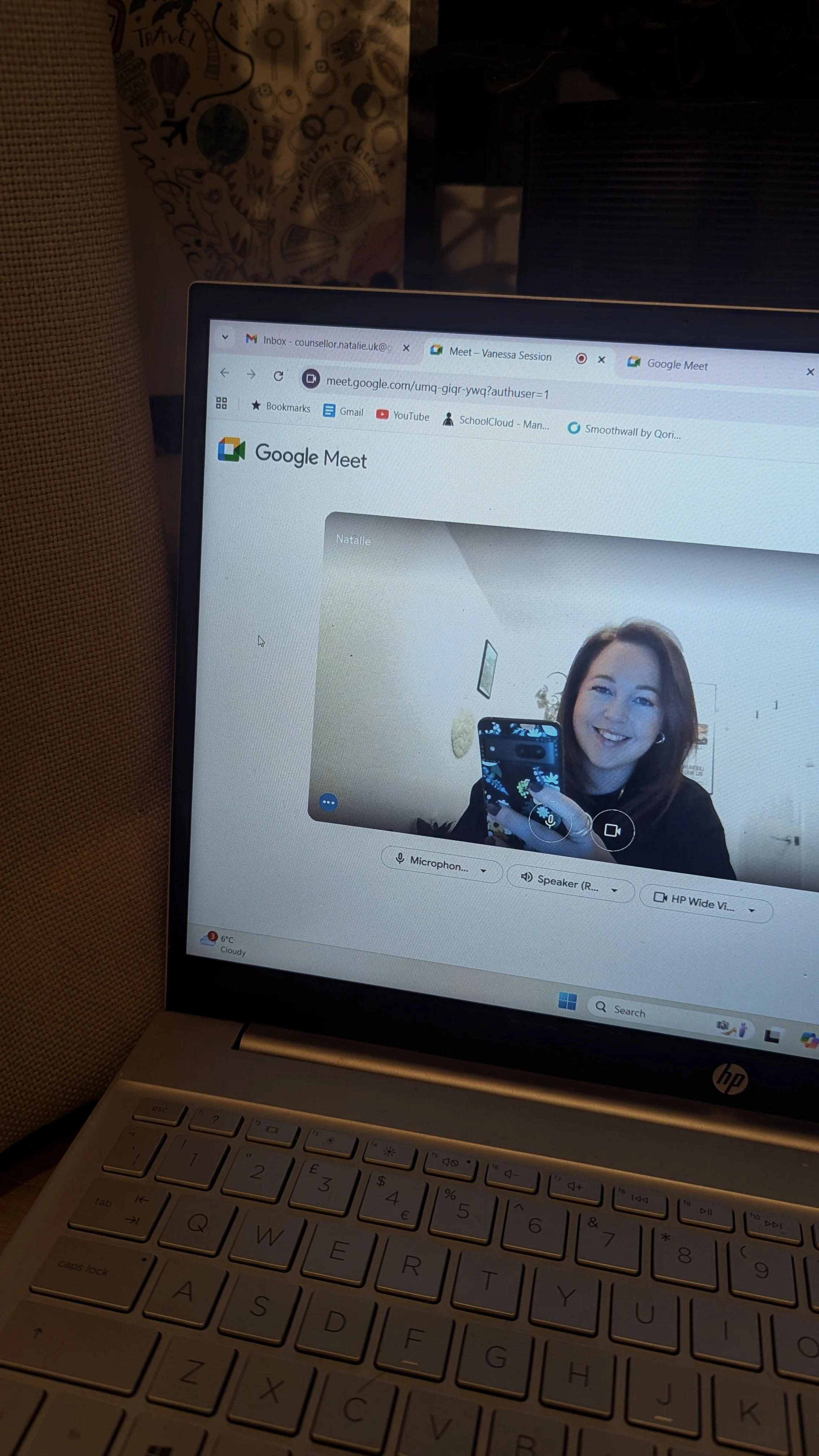 A laptop screen showing a smiling woman taking a selfie during a video call on Google Meet. The woman has shoulder-length brown hair and is holding a phone with a floral pattern case.