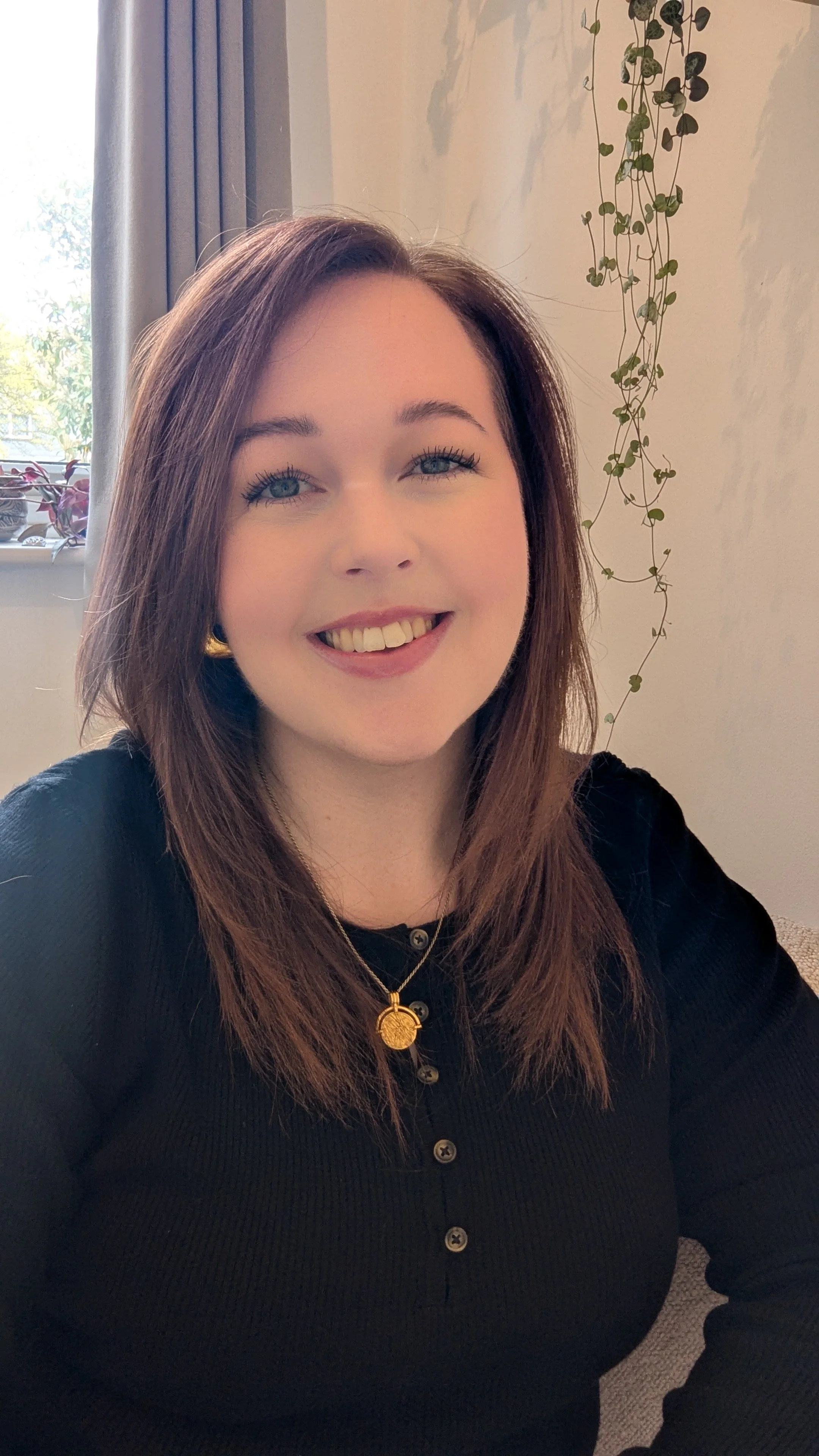 A woman with long brown hair, blue eyes, and a big smile, wearing a black top with buttons and gold jewelry, sitting indoors near a window with curtains and hanging plants in the background.