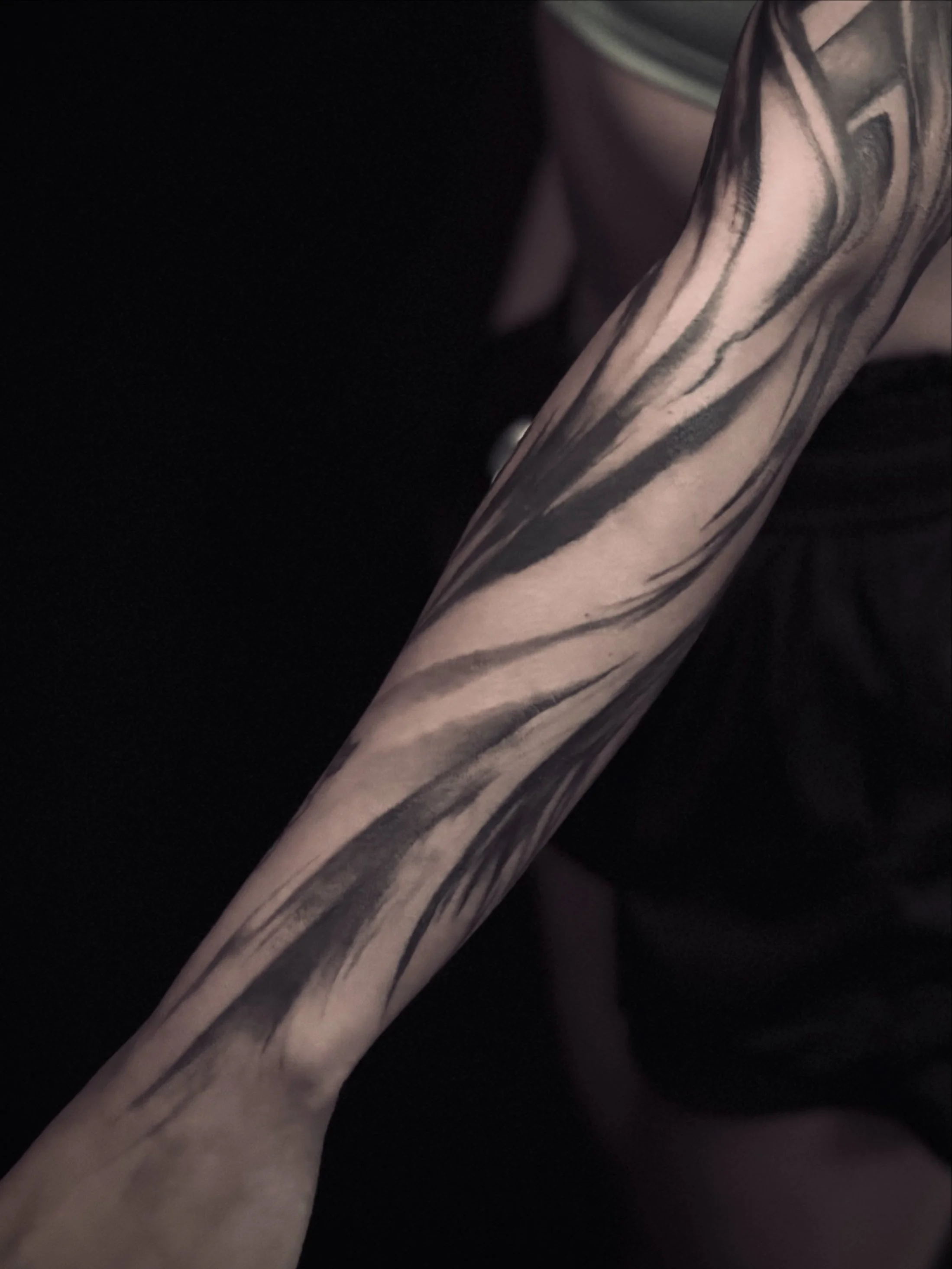 Close-up of a tattooed forearm with black ink lines and shading on light skin, against a dark background.
