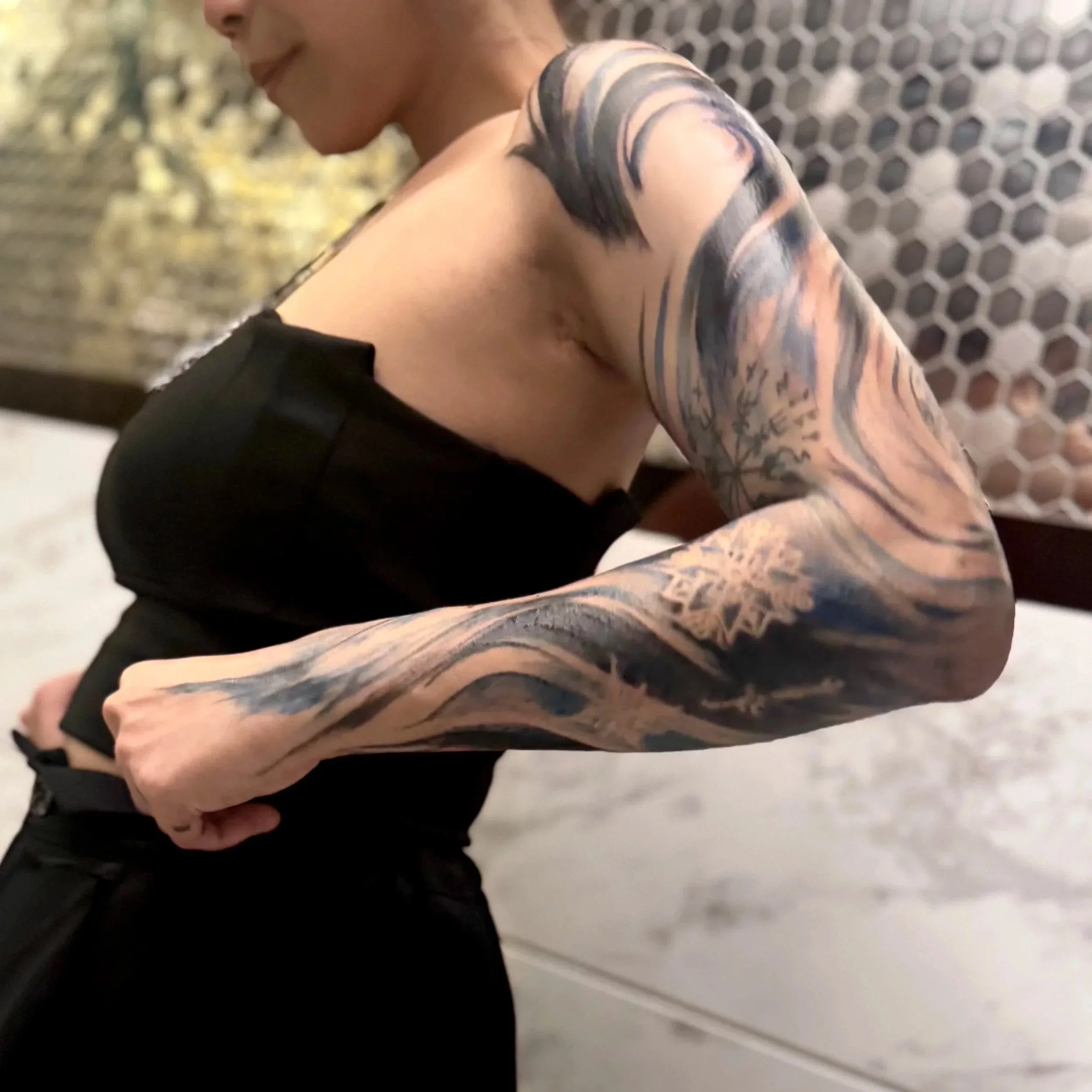 Close-up of a person's shoulder with large tattoos, wearing a short-sleeved shirt in front of a patterned wall.
