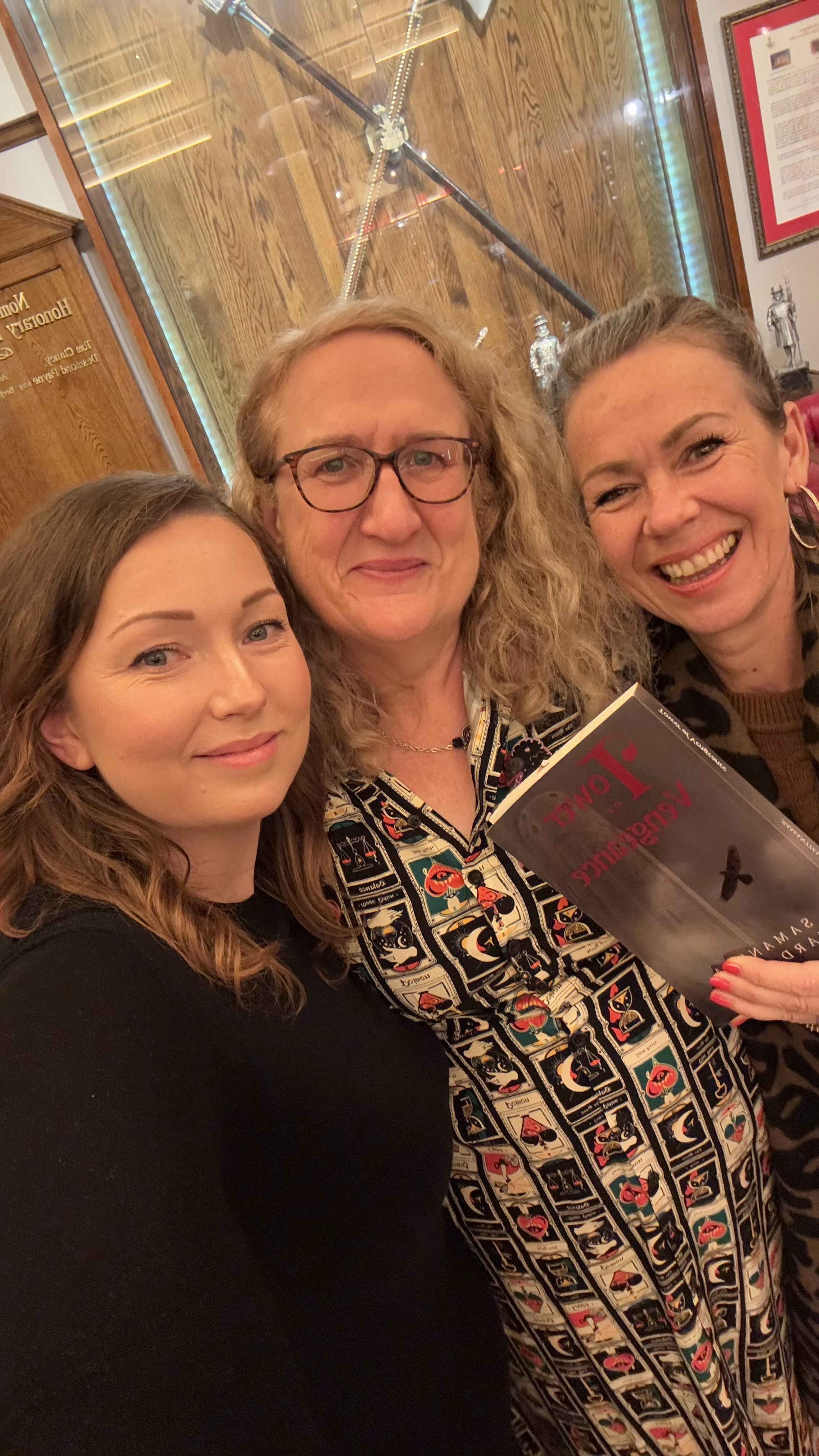 Three women taking a selfie in a museum or gallery setting with a glass display case and framed pictures on the wall behind them. One woman is holding a book titled 'Aggressively Loyal' by Samay.