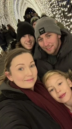 A family of four taking a selfie at a festive event with bright, decorative lights in the background.