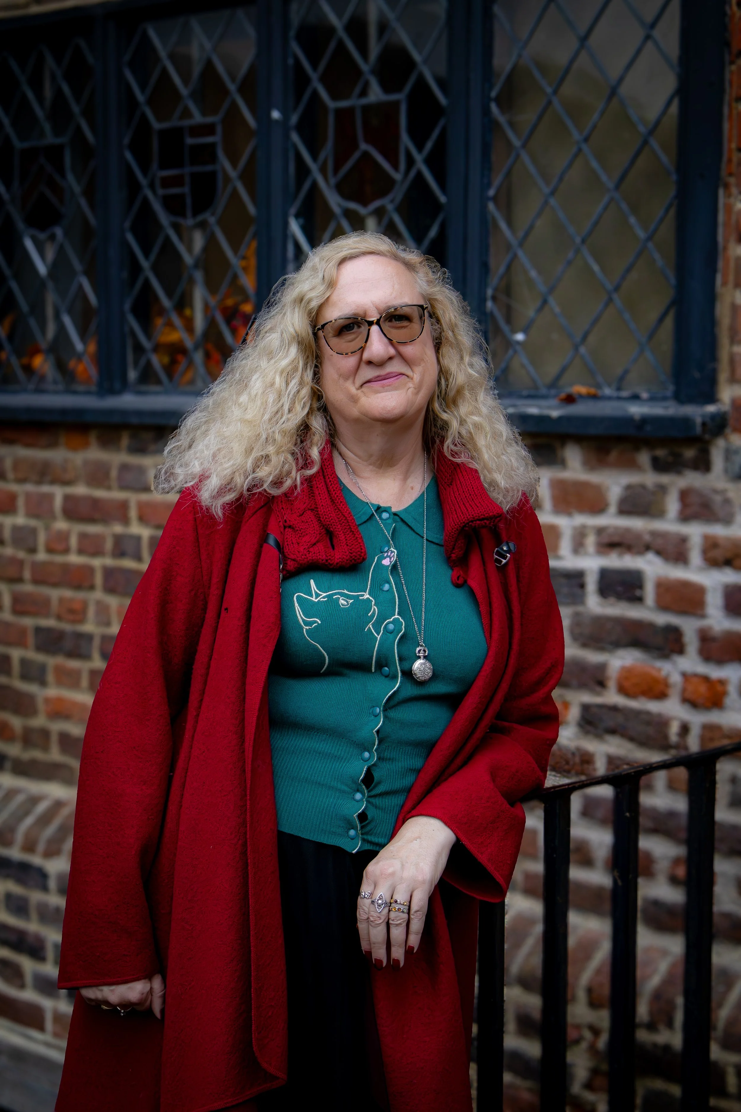 A woman with curly blonde hair and glasses, wearing a teal shirt with a cat design, a red coat, and jewelry, standing outdoors in front of a brick wall with barred windows.