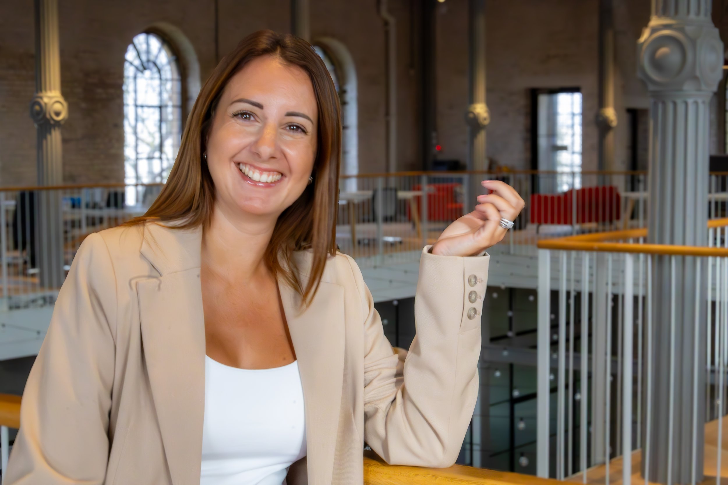 A smiling woman with shoulder-length brown hair in a beige blazer sitting in an indoor space with high windows, columns, and industrial-style decor.