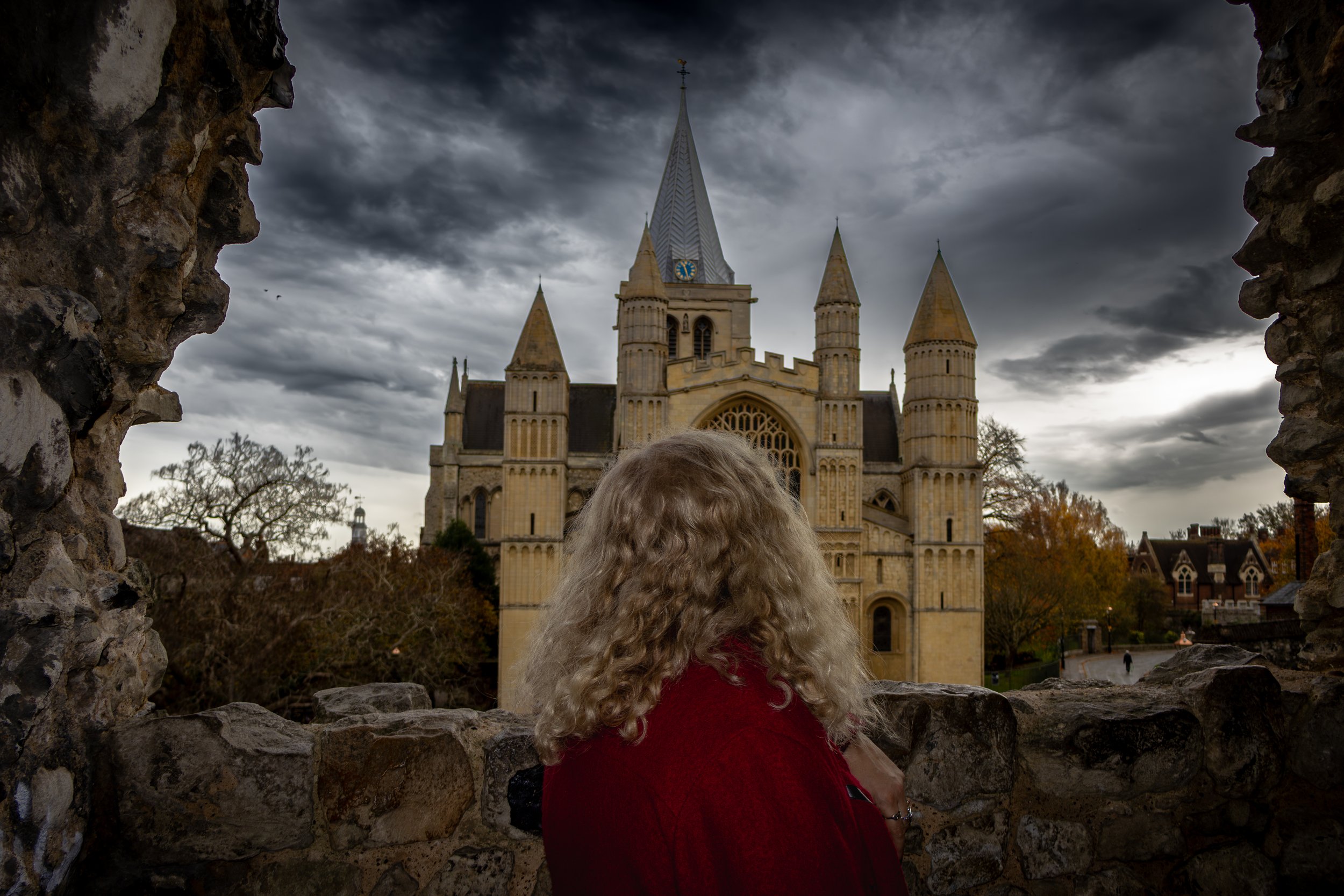 A woman with curly blonde hair, wearing a red coat, looking at a Gothic-style castle with a tall, pointed spire and four smaller towers, under a cloudy sky, viewed from behind through a stone window or opening.