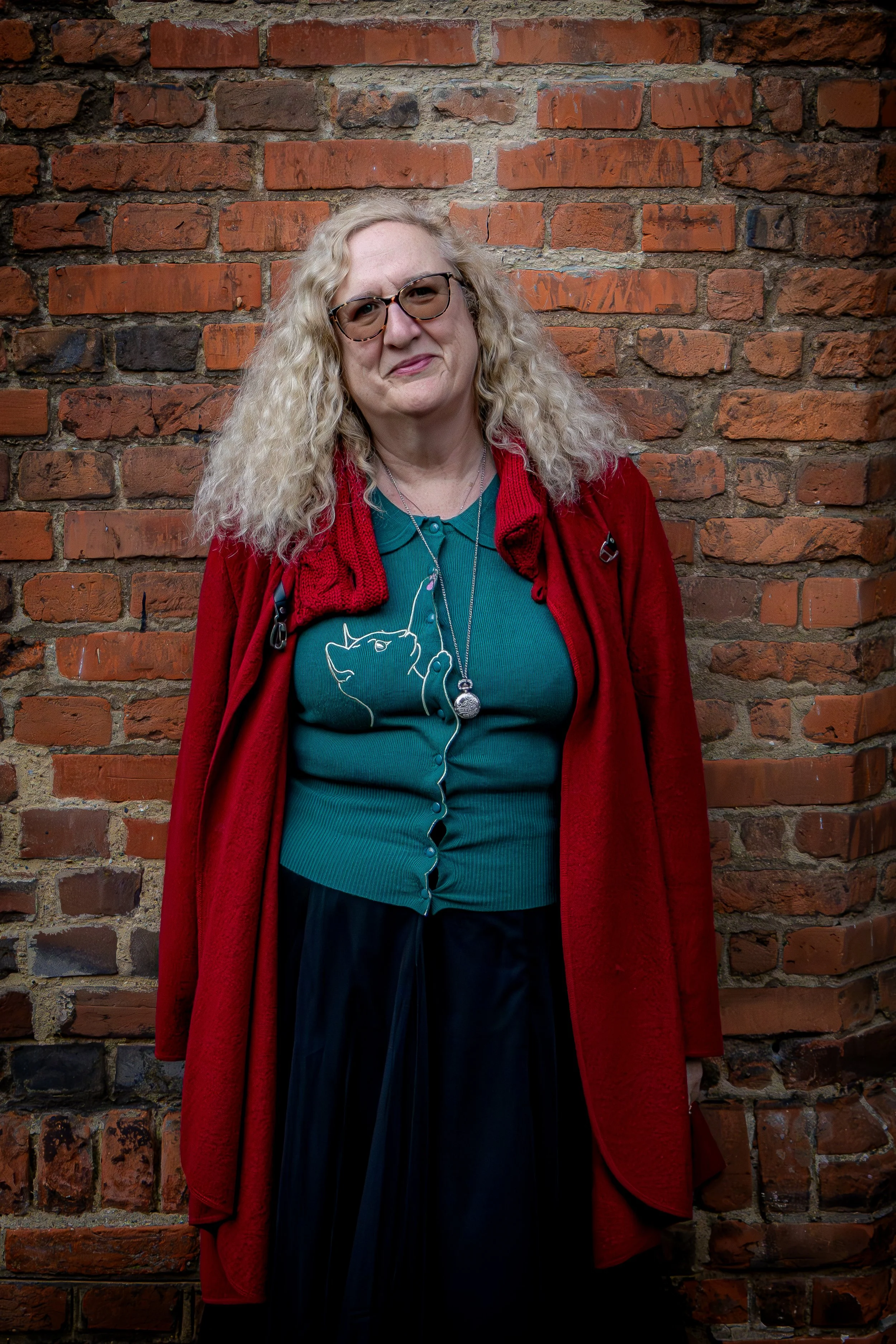 A woman with curly blonde hair and glasses standing in front of a brick wall, wearing a teal shirt, a red coat, and a necklace.