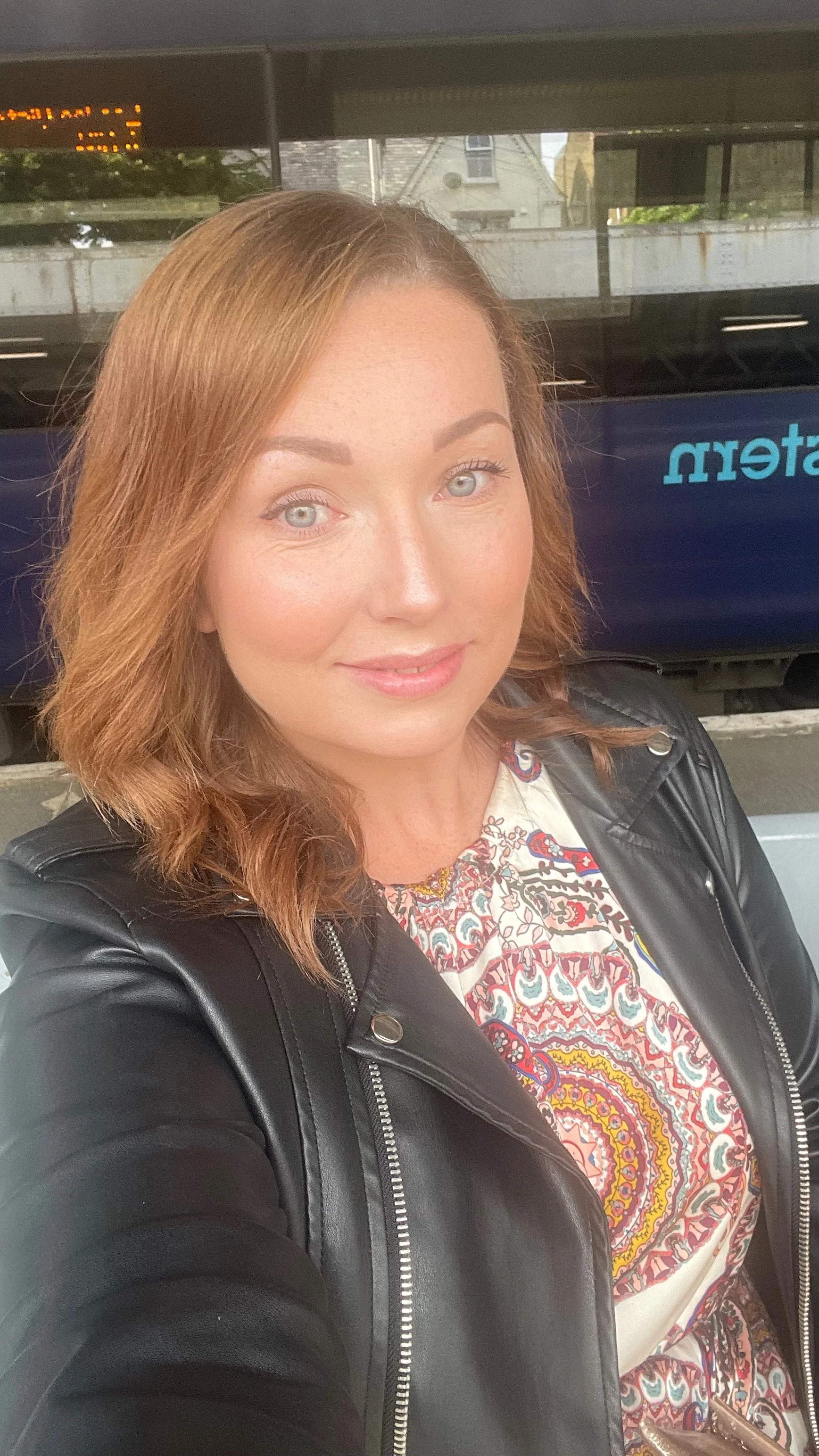 A woman with shoulder-length red hair, wearing a black leather jacket and a colorful patterned top, taking a selfie at a transportation station.