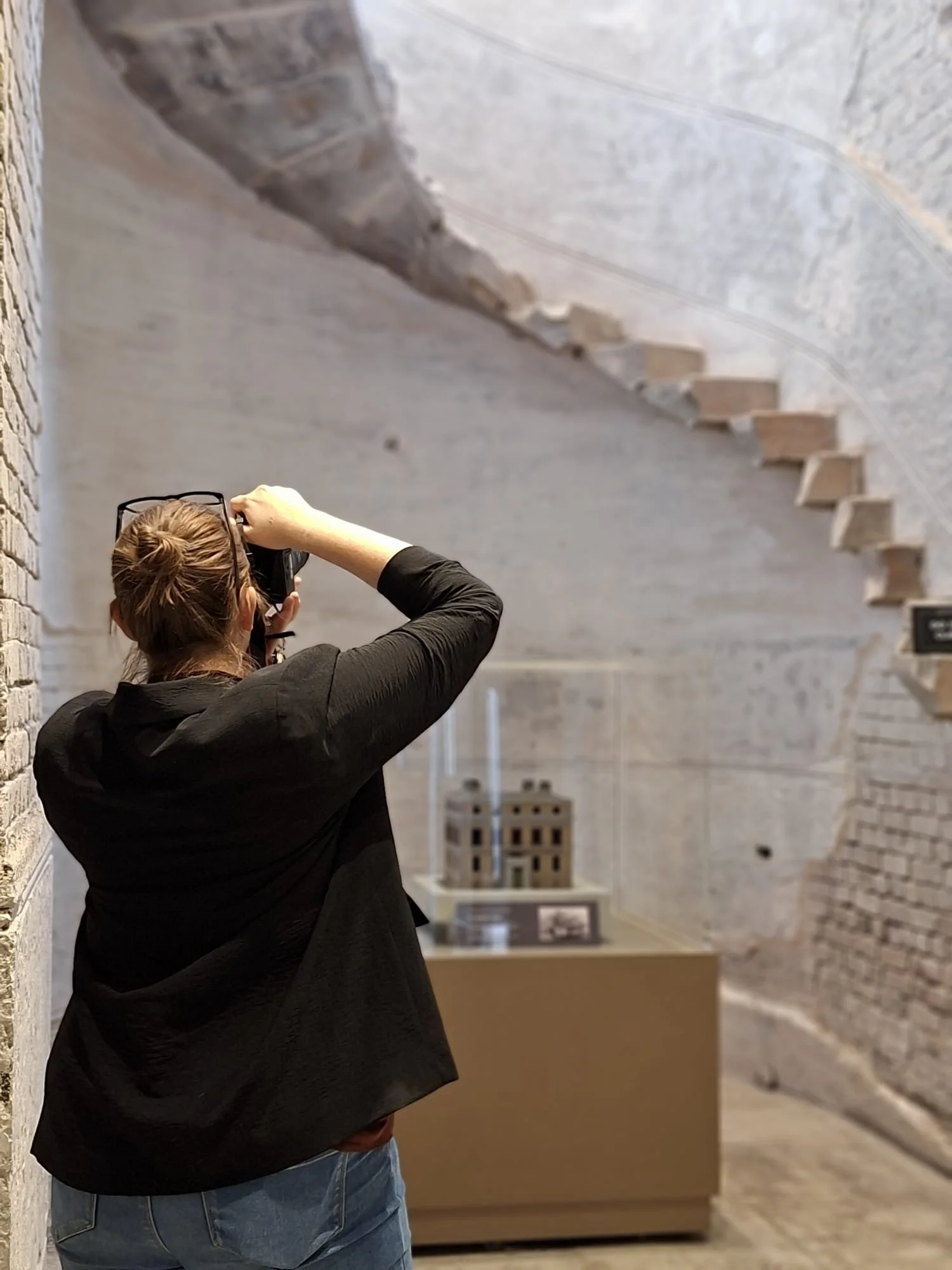 A woman with glasses on her head taking a photo of a model building in a museum or exhibit with rough brick and concrete walls and a spiral staircase in the background.