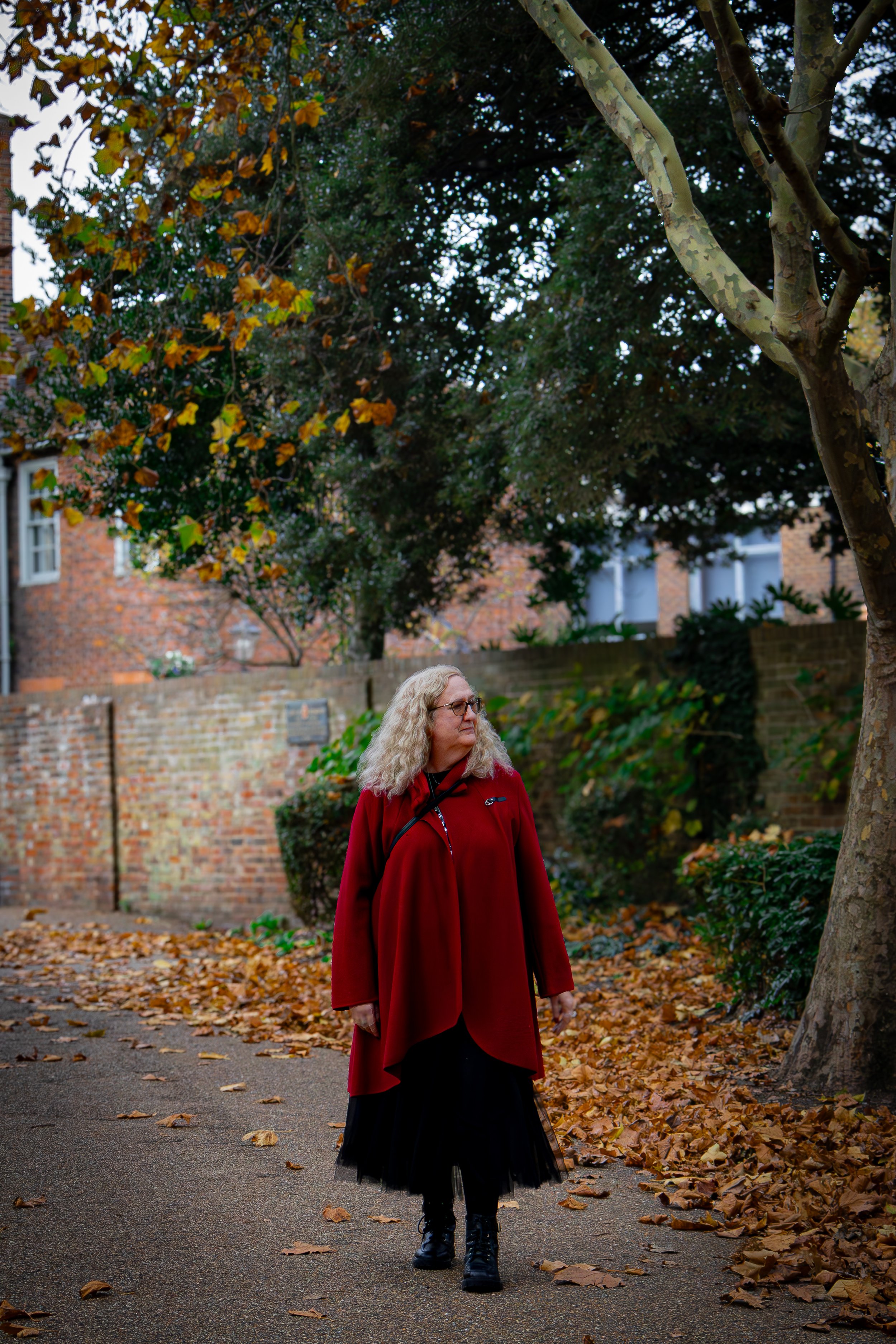 A woman with curly blonde hair, wearing glasses, a red coat, and black boots, walking on a leaf-covered path in a park during autumn.