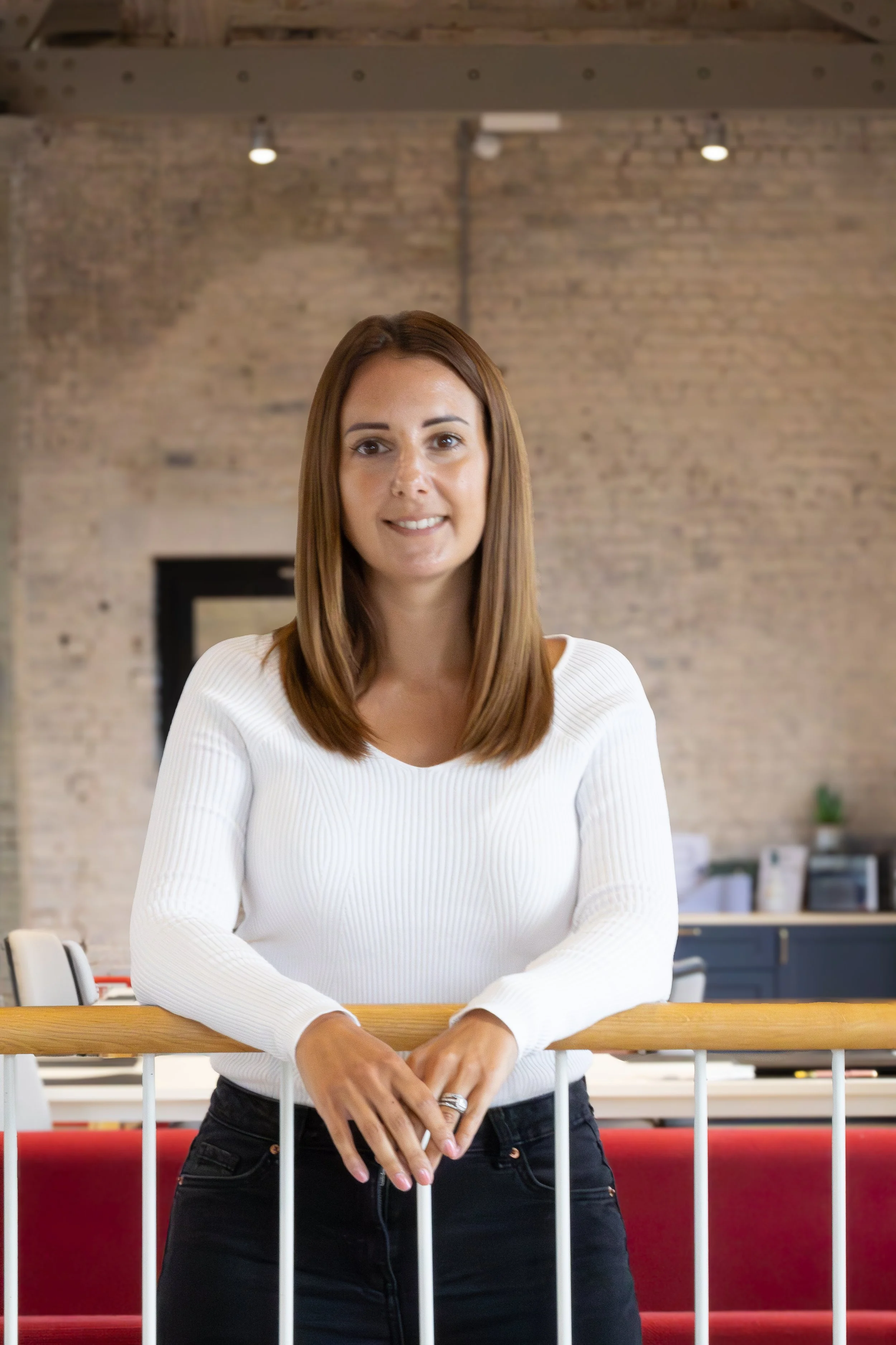 A woman with medium-length brown hair, wearing a white long-sleeve top, standing indoors behind a wooden railing, with an exposed brick wall and some furniture in the background.
