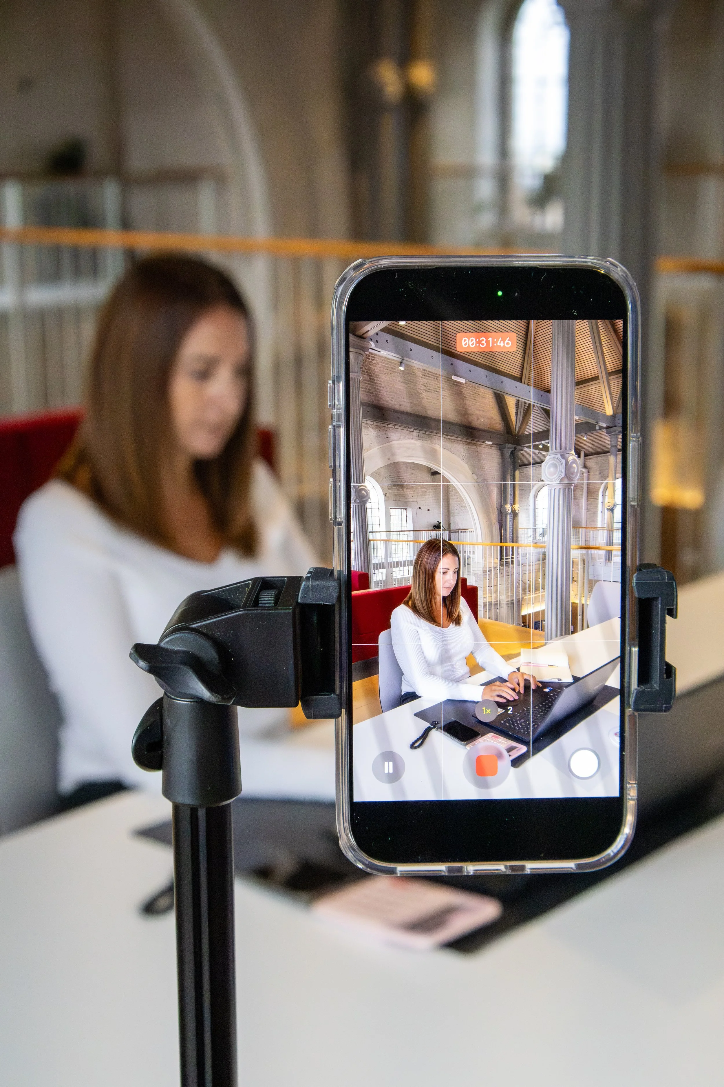 A woman sitting at a desk working on a laptop, with her image being recorded by a smartphone mounted on a tripod in front of her, capturing her in a recording studio or office environment.
