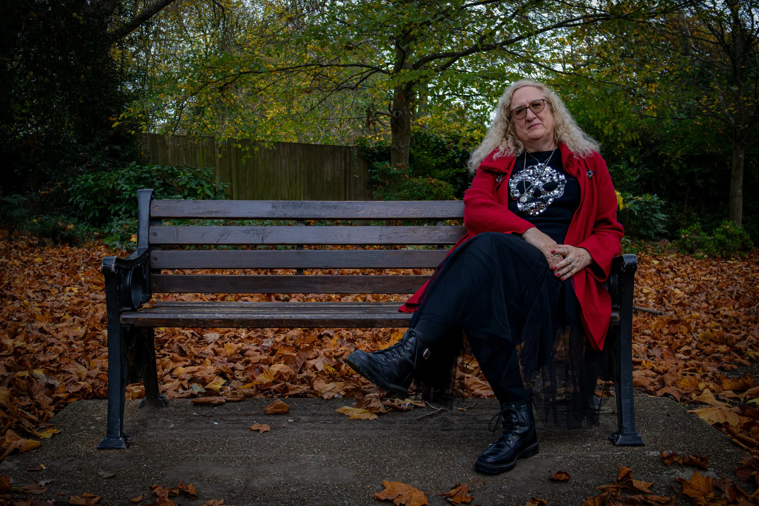 A woman with curly blonde hair, glasses, wearing a red coat, black t-shirt with a skull design, black skirt, and black combat boots, sitting on a park bench in an autumn scene with fallen leaves around.