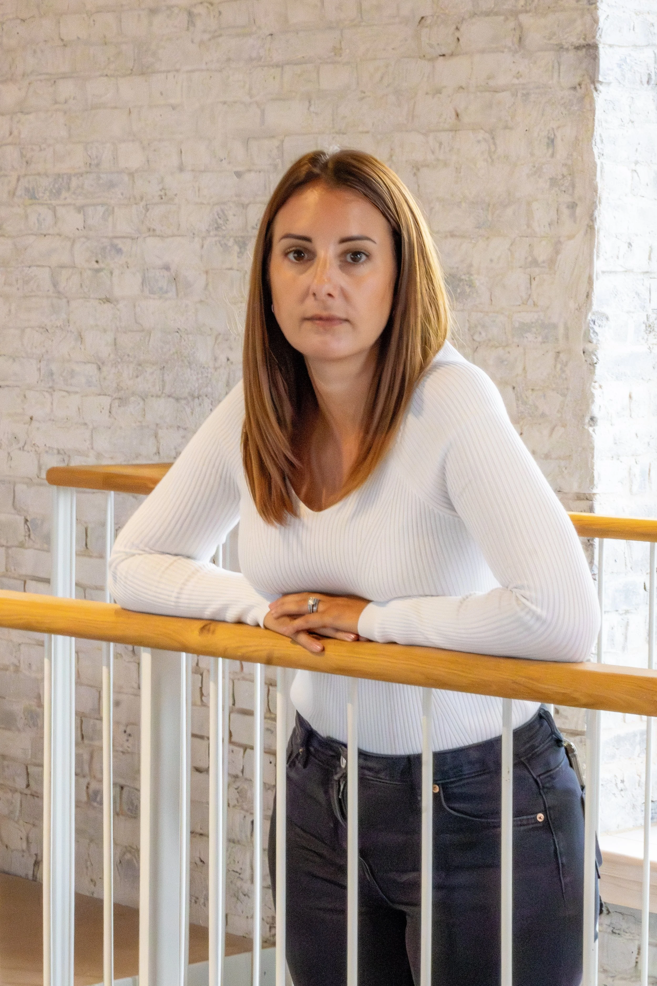 A woman with shoulder-length brown hair, wearing a white long-sleeve top and dark jeans, leaning on a wooden railing in front of a white brick wall.