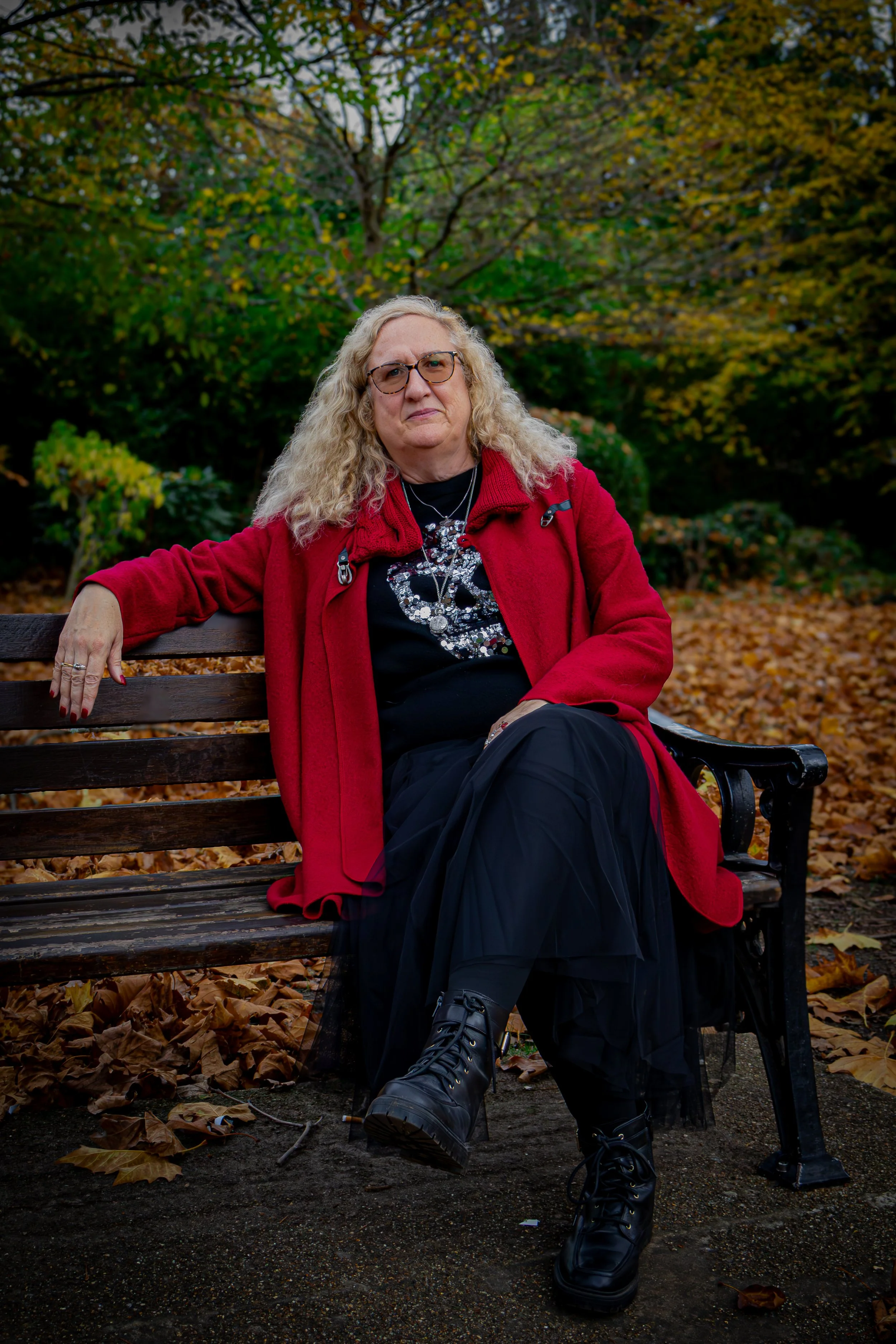 Older woman with curly blonde hair and glasses sitting on a wooden park bench in fall, wearing a red coat, black t-shirt with a heart design, a long black skirt, and black boots, with autumn leaves and trees in the background.