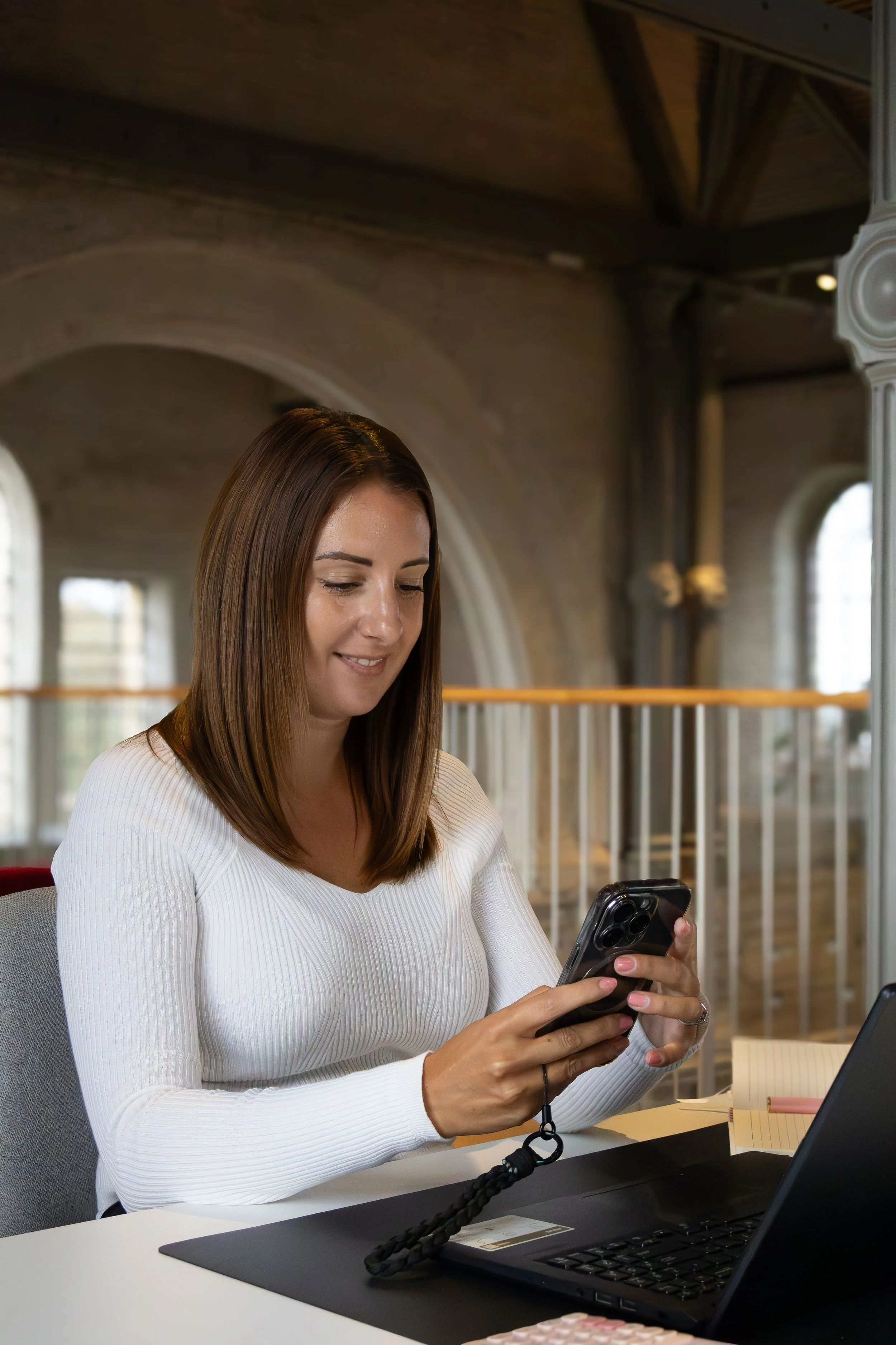 Woman with shoulder-length brown hair using smartphone, sitting at a desk with a laptop, notebook, and pen in a room with large windows and wooden beams.