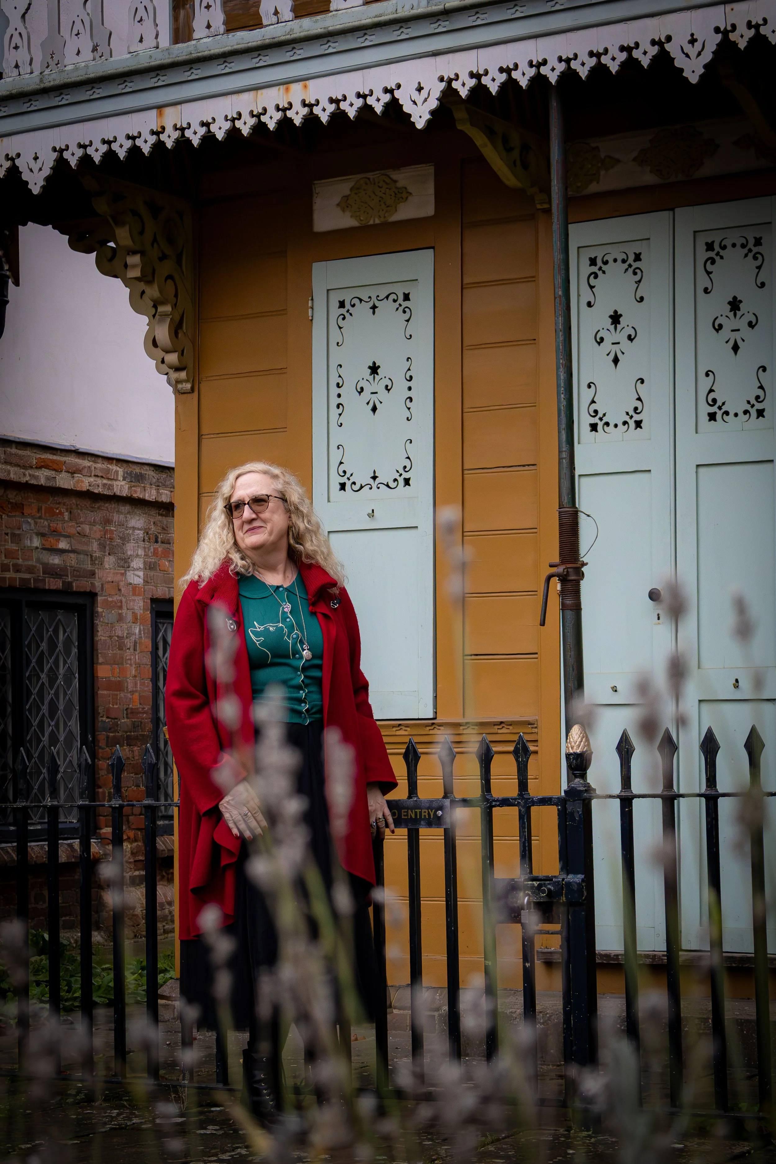 A woman with blonde curly hair, wearing glasses, a teal shirt, a red coat, and beige gloves, standing outside a house with intricate wooden trim and pastel blue shutters.
