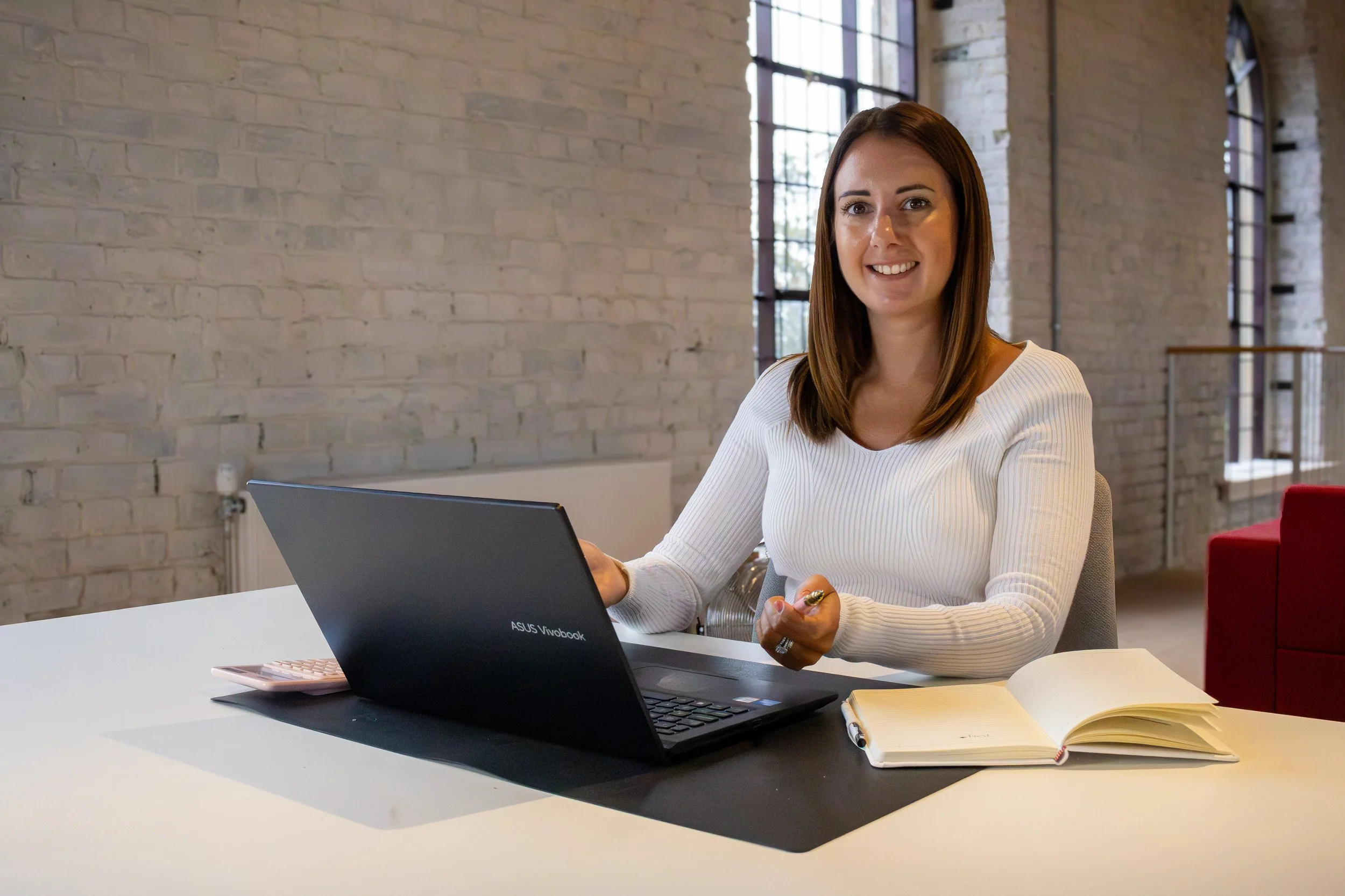 A woman with brown hair wearing a white top, sitting at a desk with a laptop, notebook, and pen in a bright office with brick walls and large windows.