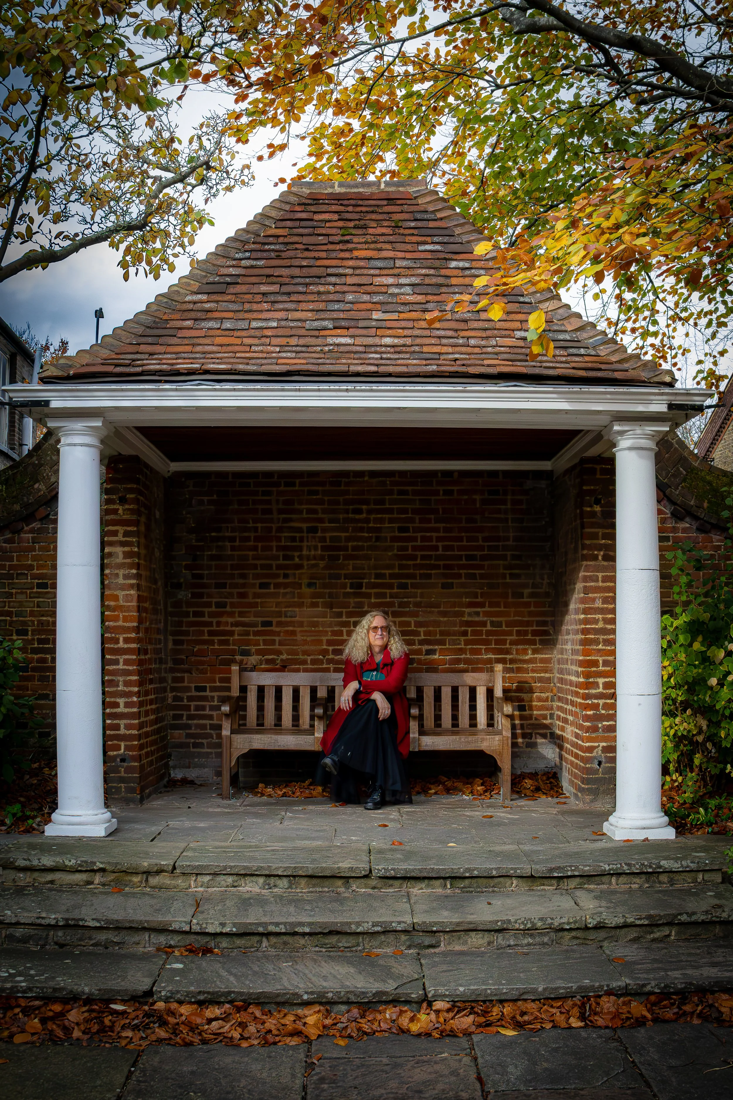 A woman with curly blonde hair and glasses sitting on a wooden bench in a small brick pavilion with a red-tiled roof, surrounded by autumn foliage.