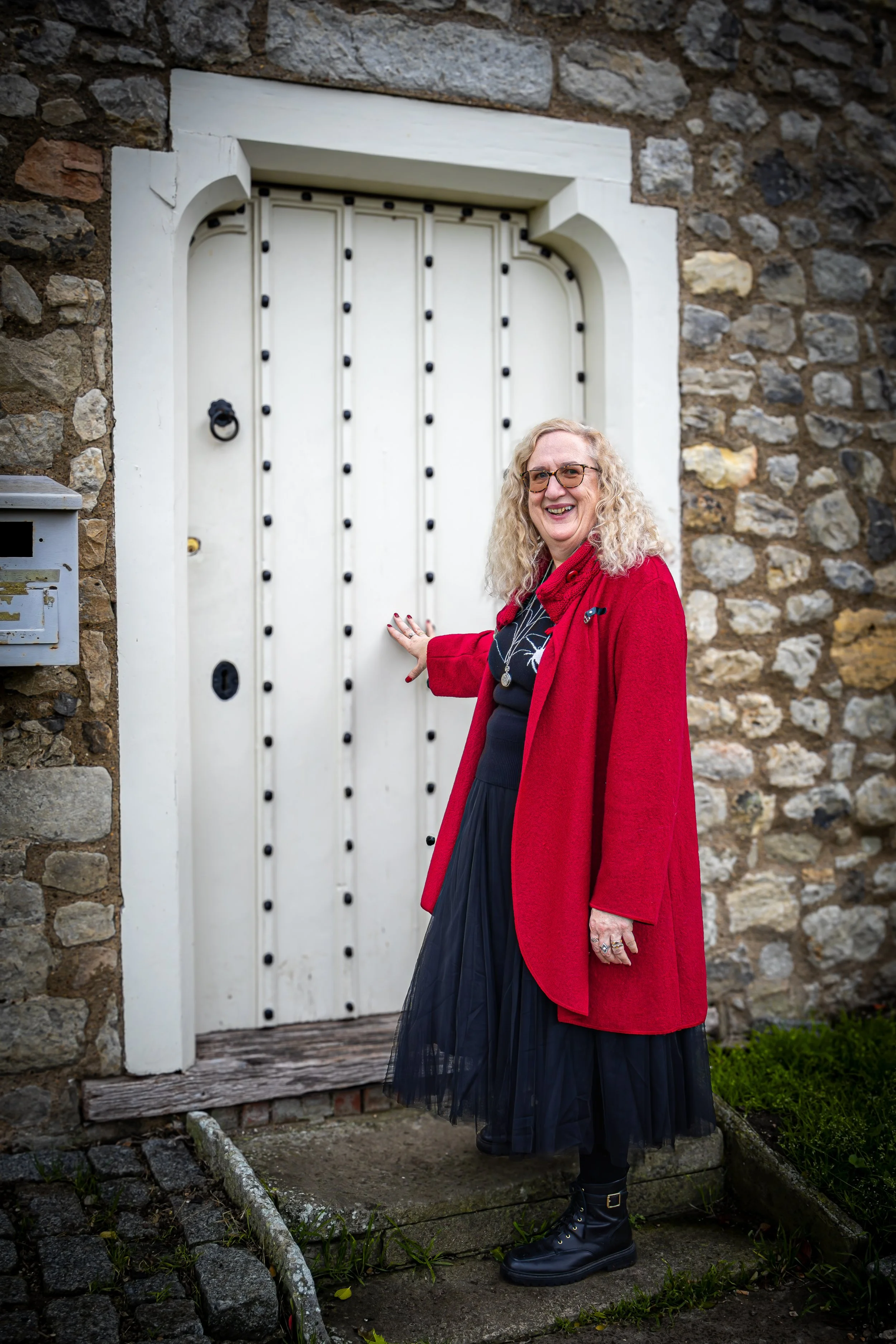 A woman with curly blonde hair and glasses standing in front of a white wooden door set into a stone wall, smiling and wearing a red coat, black dress, and black boots.