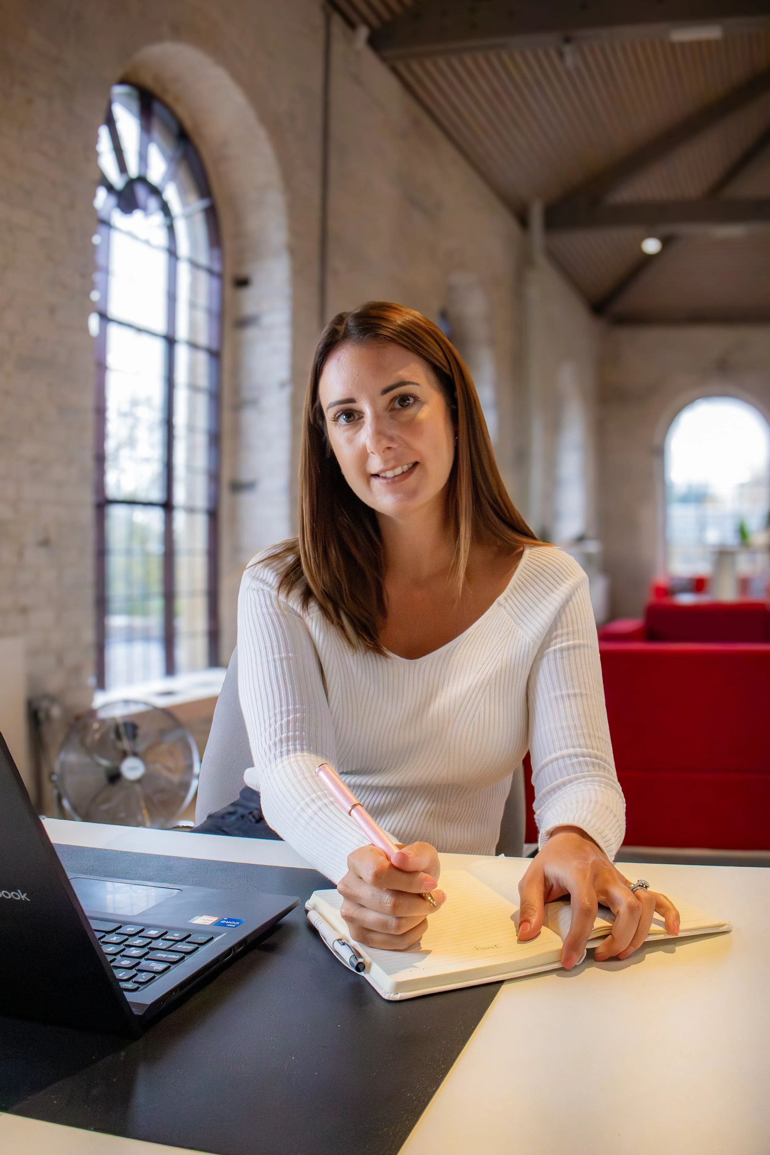 A woman with brown hair wearing a white long sleeve shirt sitting at a desk with an open laptop, writing in a notebook with a pink pen in a modern, loft-style office space with large arched windows and red furniture.