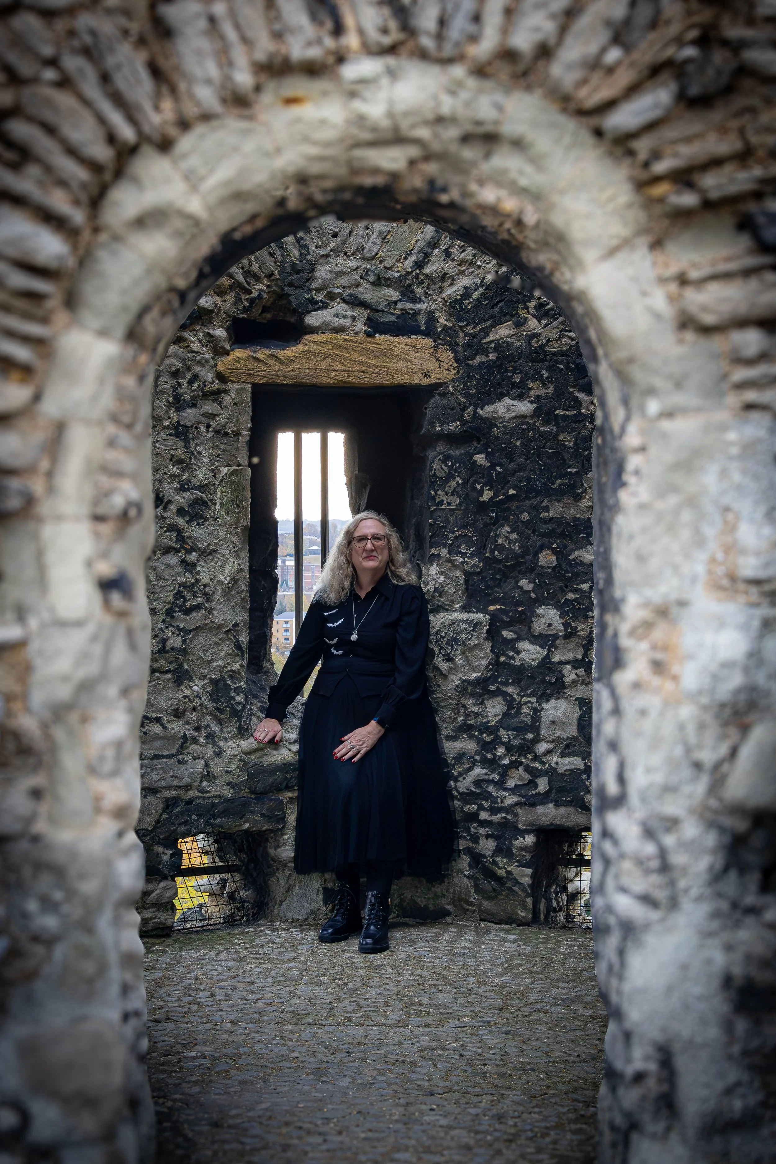 A woman wearing a black dress and boots standing inside an old stone castle tower, looking through a small barred window with a view of the cityscape outside.